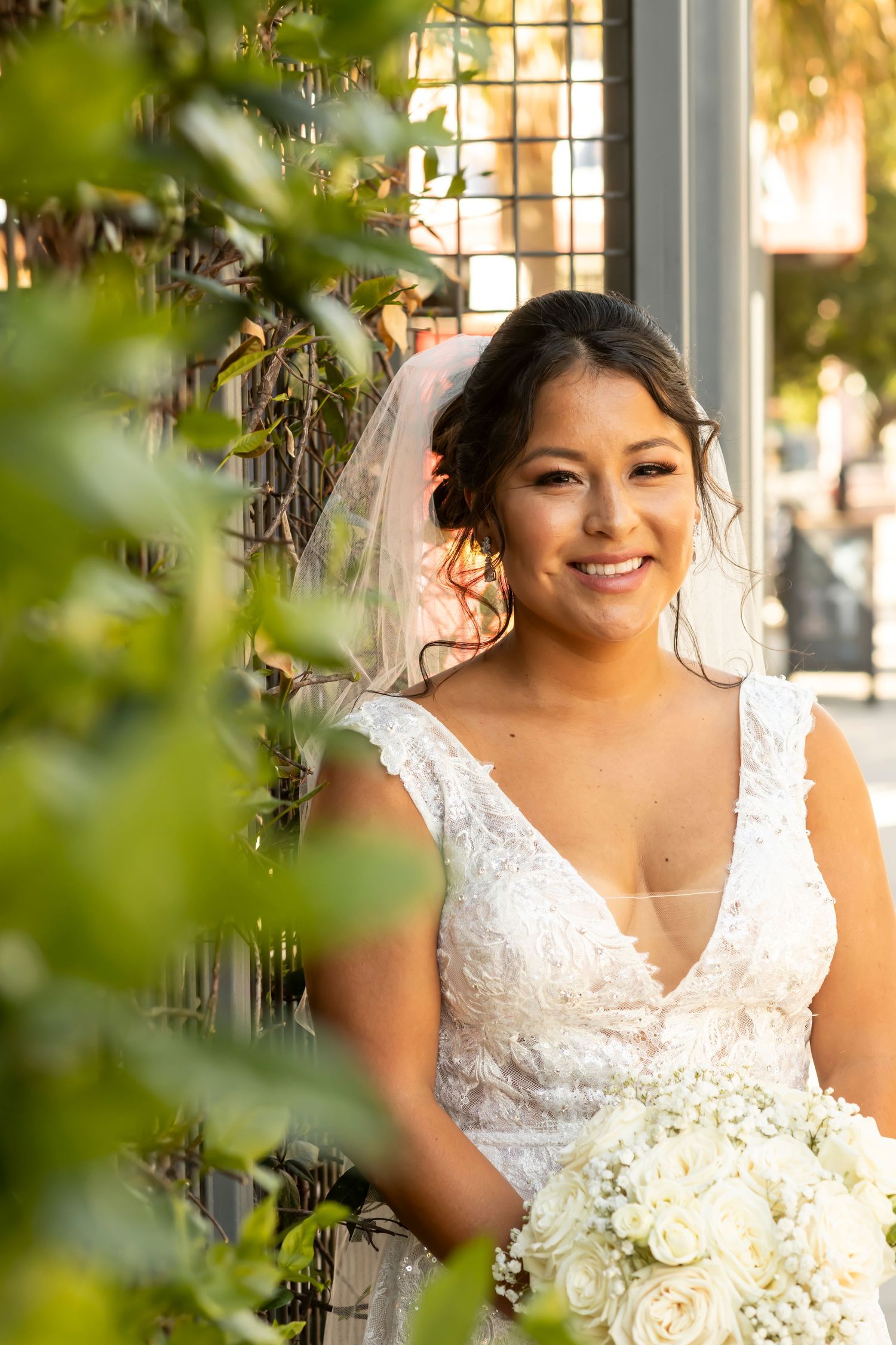 A bride in a white lace V-neck wedding dress holds a white rose bouquet while standing among green foliage.
