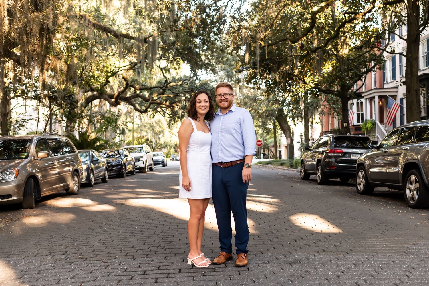 A couple stands together on a cobblestone street lined with parked cars and autumn trees casting dappled shadows.