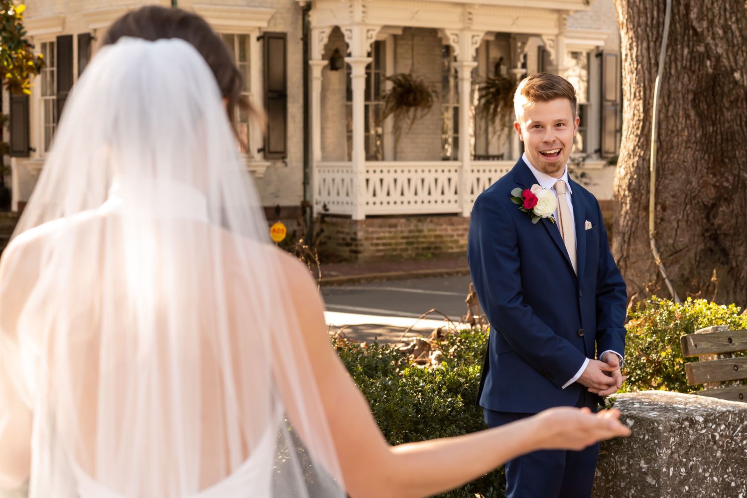 A happy groom in a navy suit and boutonniere reacts to seeing bride in her veil during their first look at a Victorian venue.