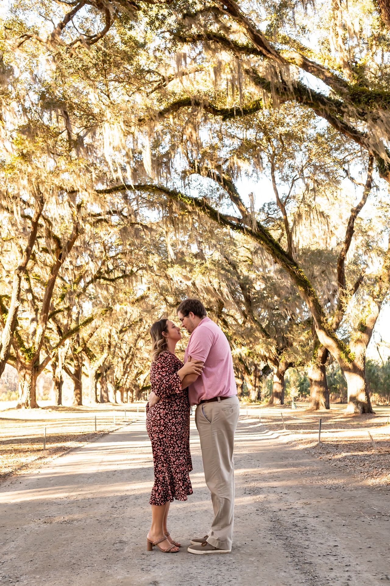A couple shares a romantic moment beneath Spanish moss-draped oak trees on a sunlit dirt path.