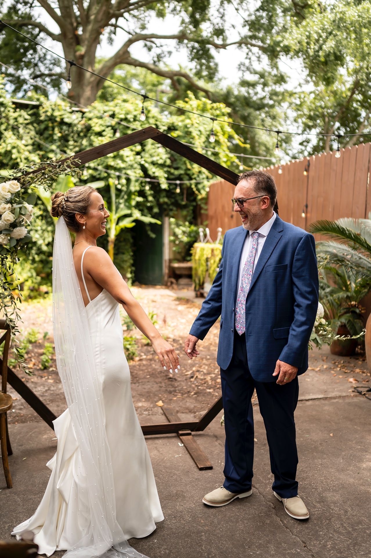 A couple shares a joyful moment in their wedding attire under trees in a backyard garden setting.
