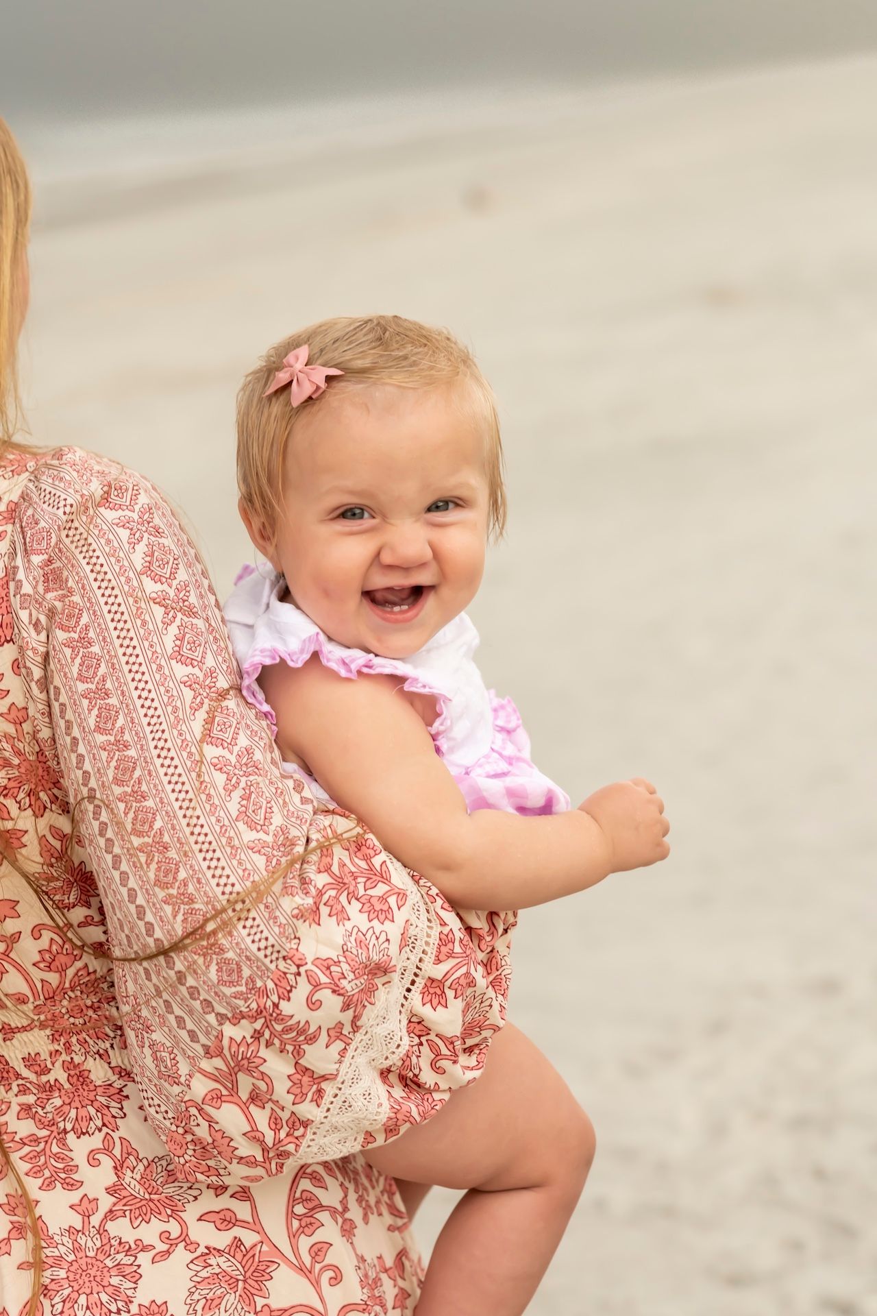 A baby wearing a pink dress sits happily against someone in a coral patterned dress on a light background.