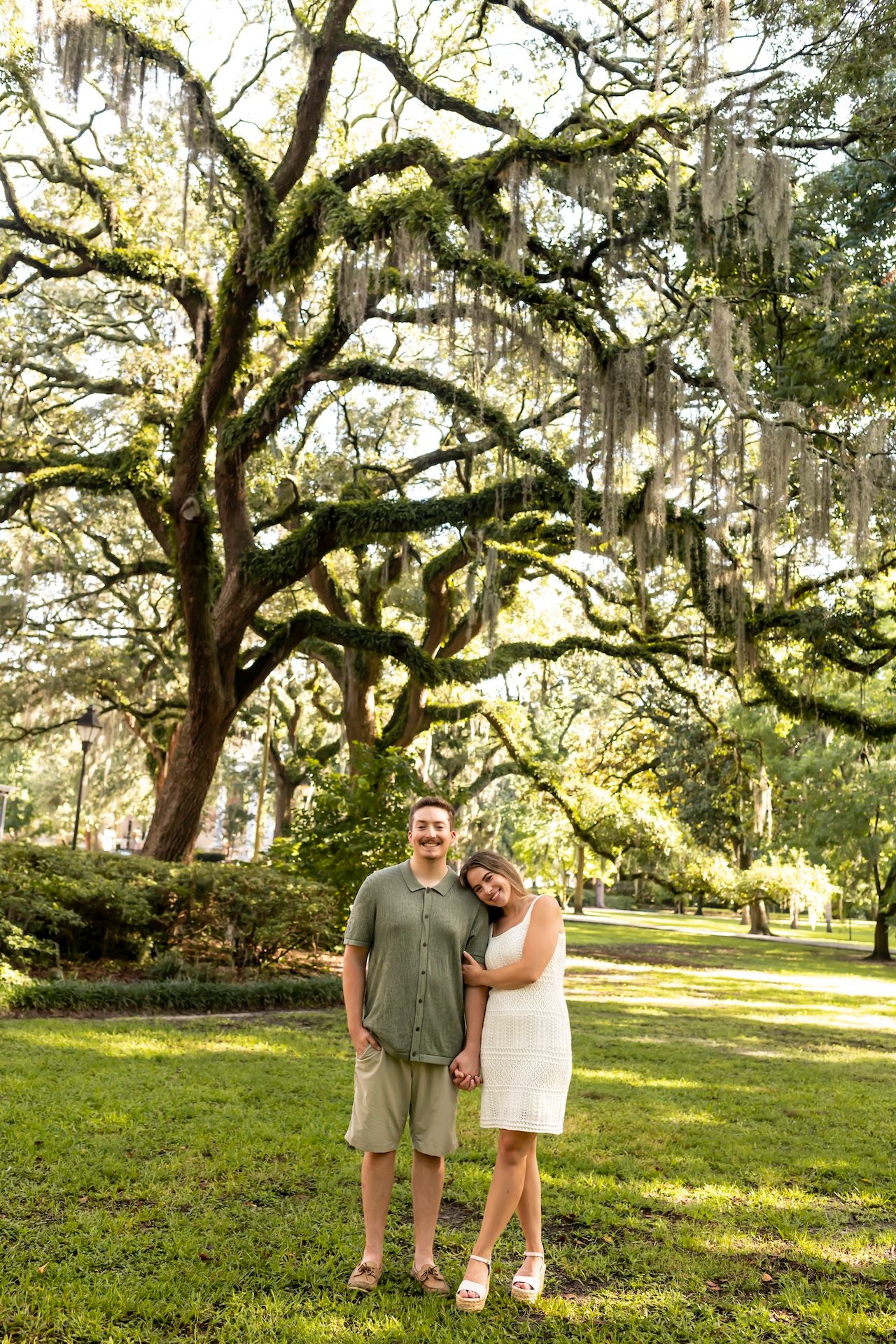 A couple stands together under a majestic live oak tree draped with Spanish moss in a serene park setting.