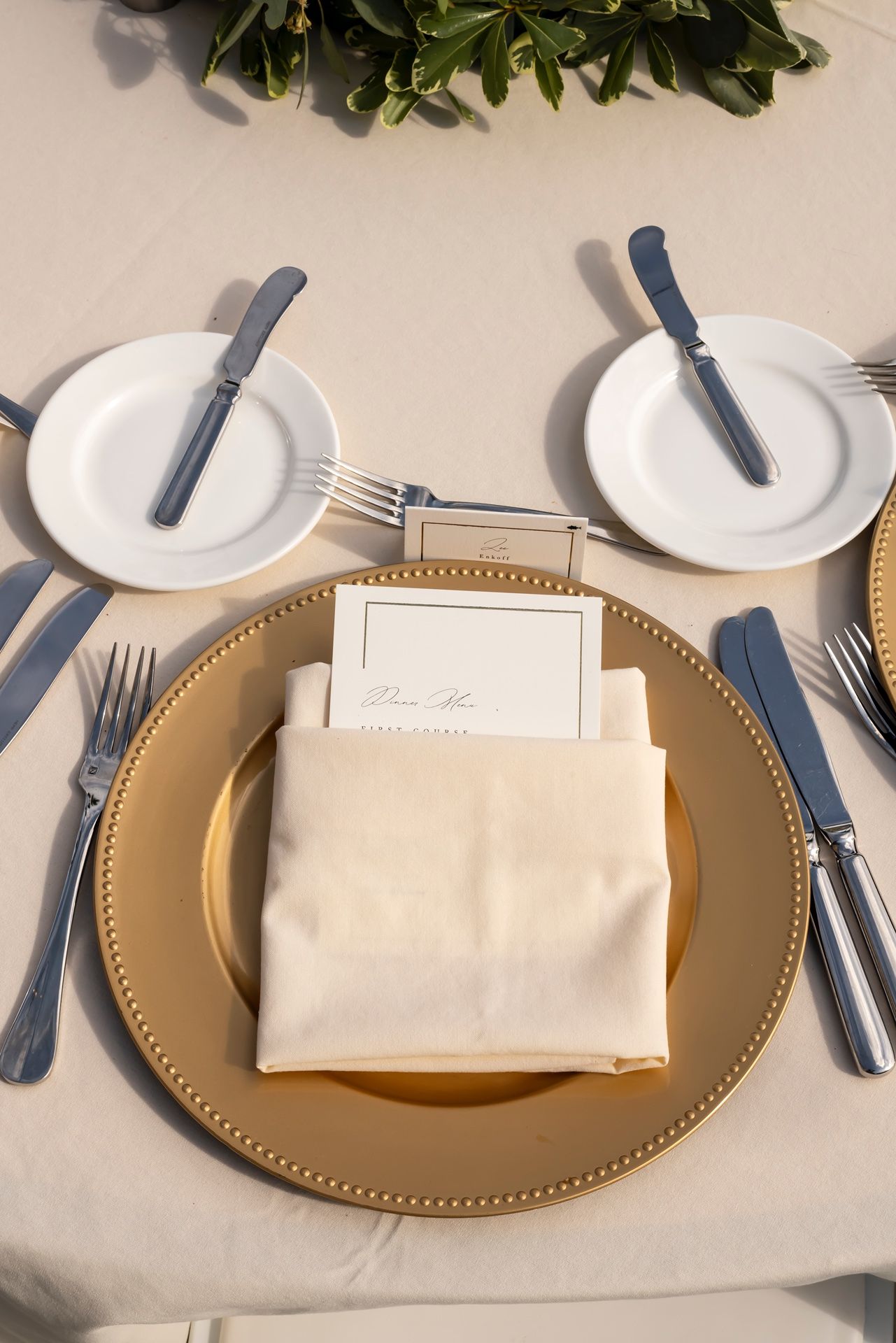 Elegant place setting with white napkin, gold charger plate, and cutlery arranged on a white tablecloth.