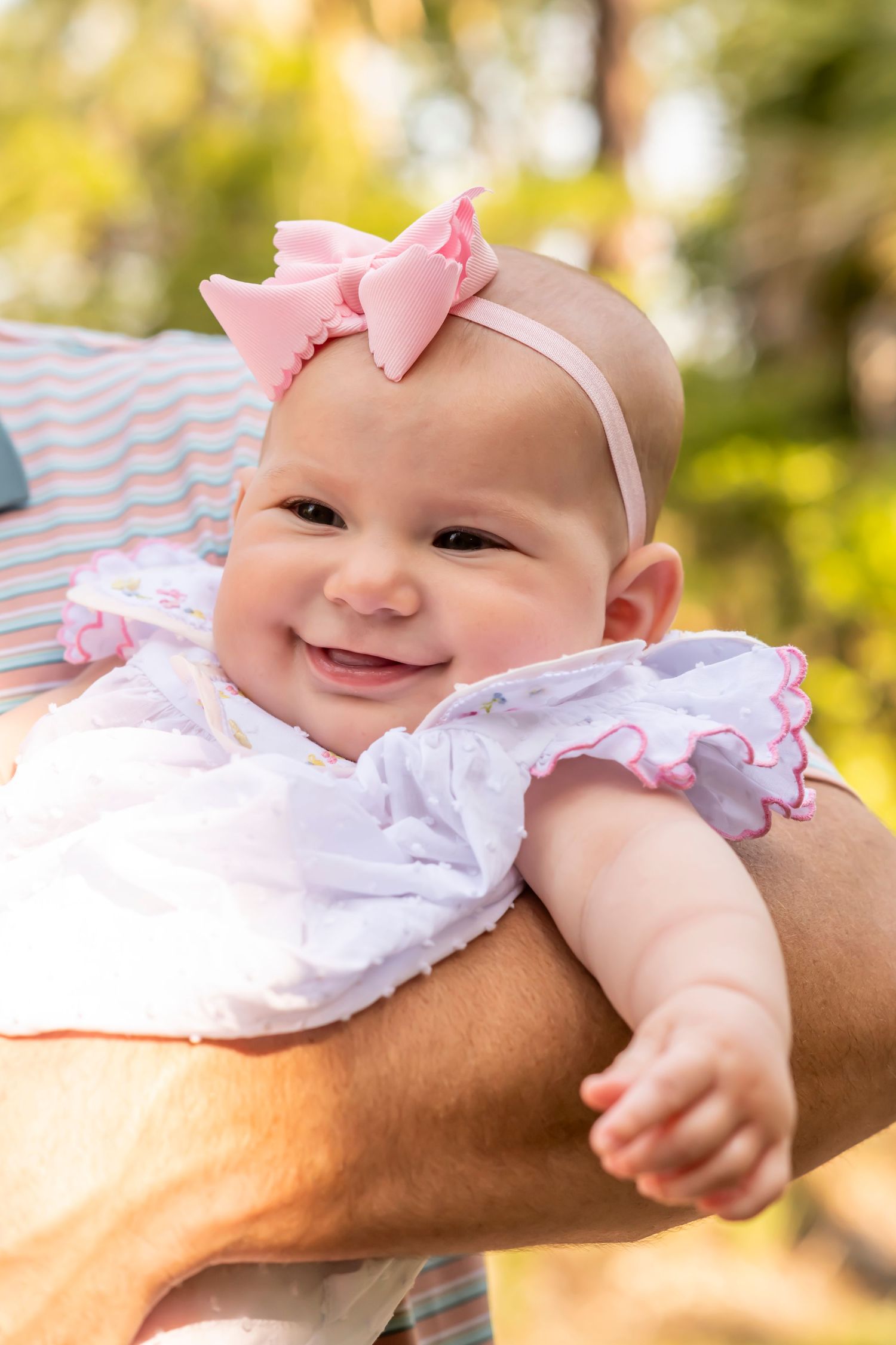 A baby in a white dress and pink bow headband smiles while being held outdoors in bright sunlight.