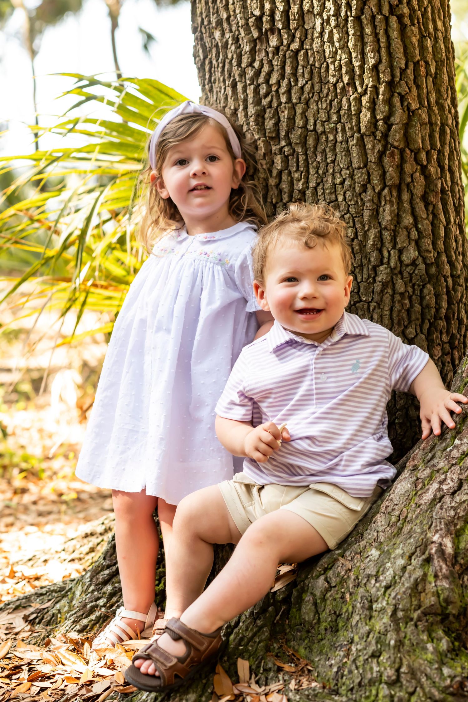 Two young children in light summer clothing sit together against a large tree trunk in dappled sunlight.