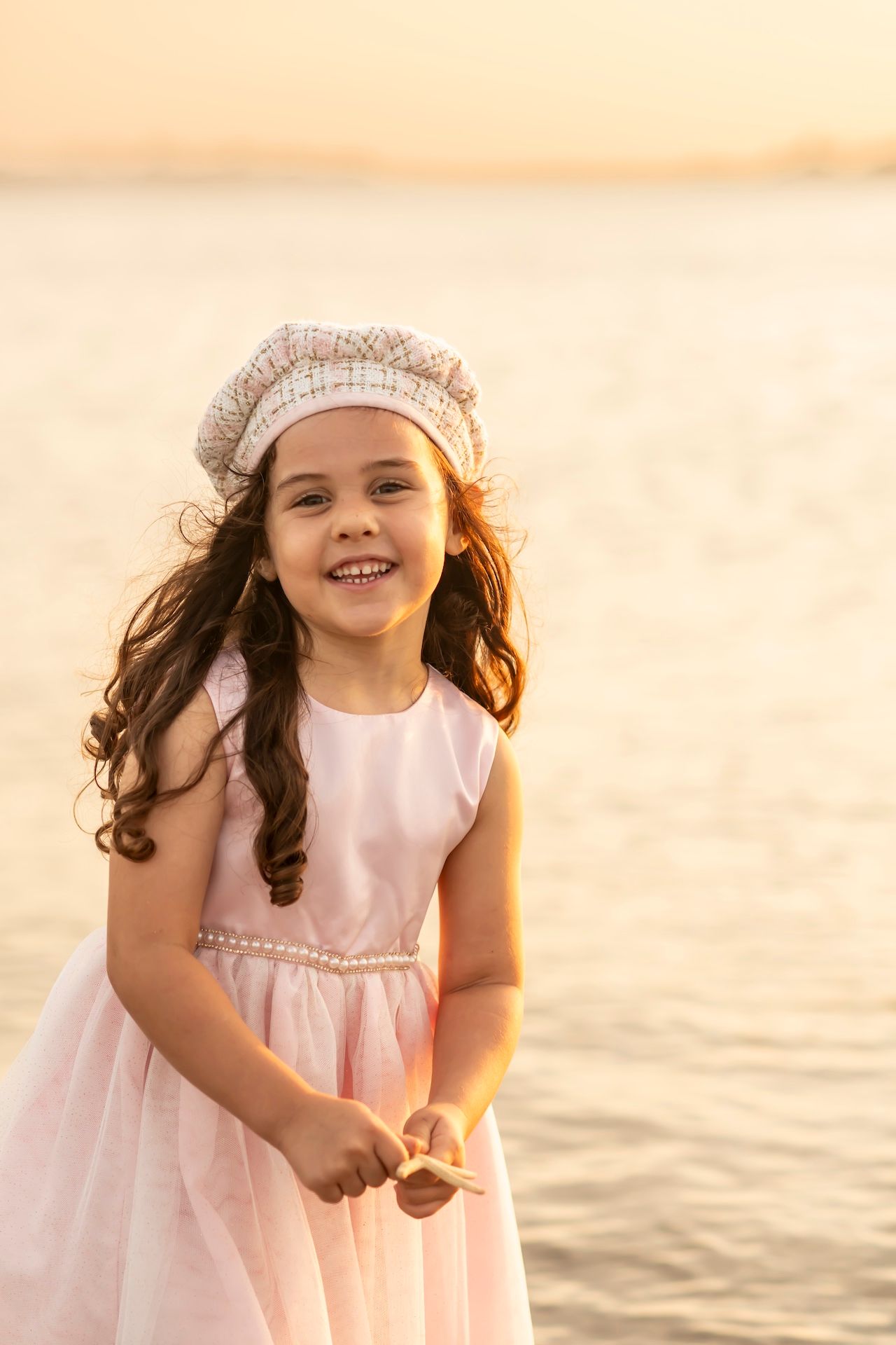 A child in a pink dress and beige hat smiles joyfully on the beach during golden hour.