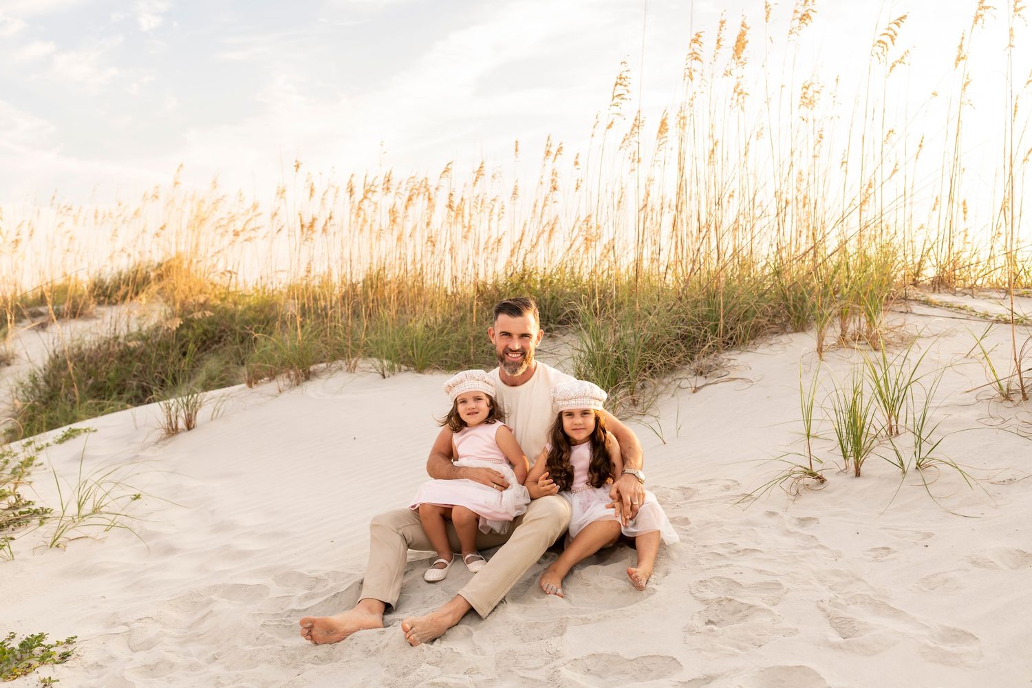 A smiling family sits together on a sandy beach dune with tall grass swaying in the golden evening light.