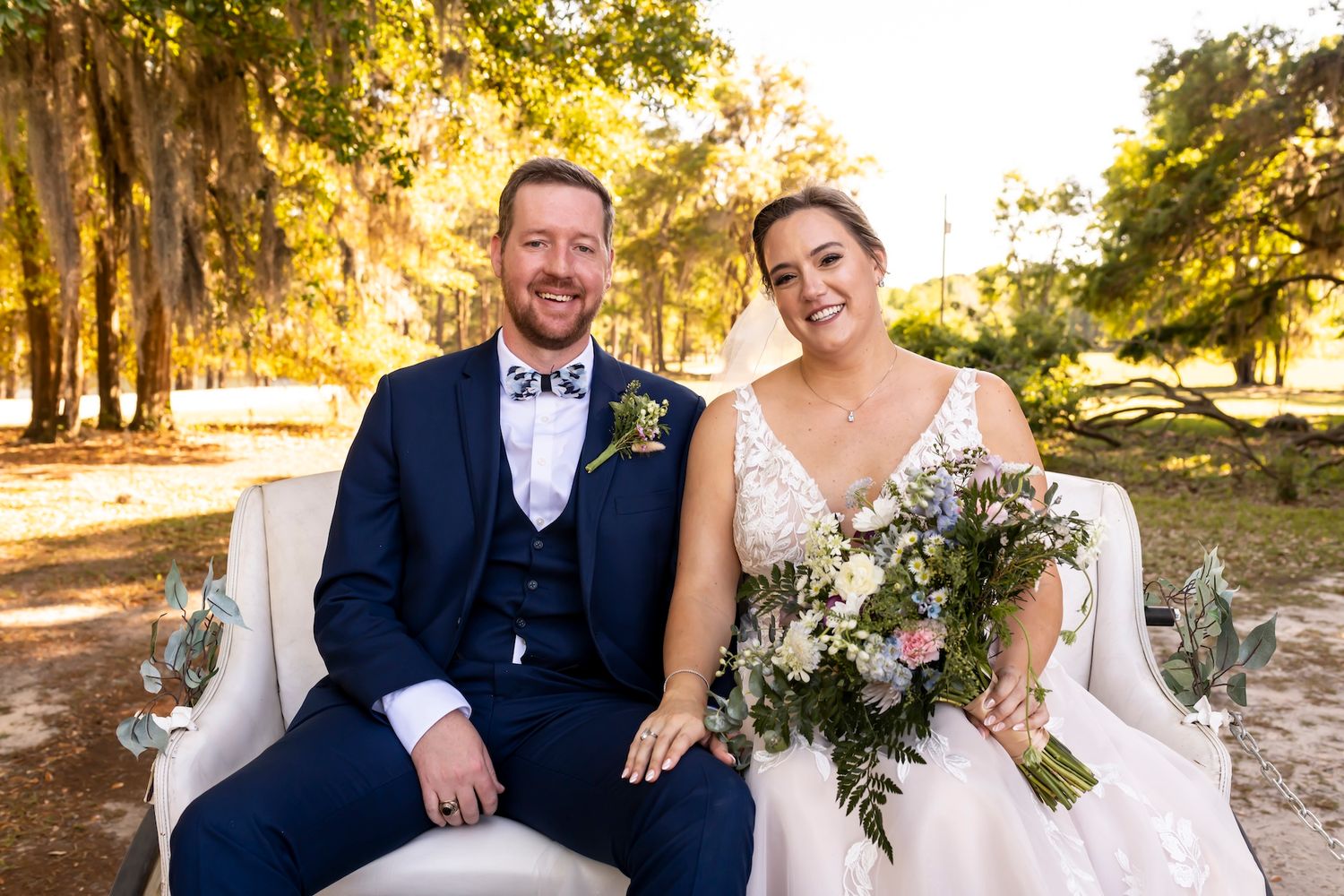 Newlywed couple sits together on vintage sofa outdoors with sunlit trees and Spanish moss in background.