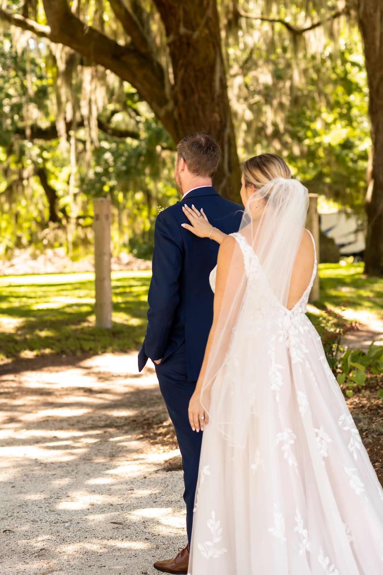 A couple in wedding attire stands on a sunlit path surrounded by trees with dangling moss.