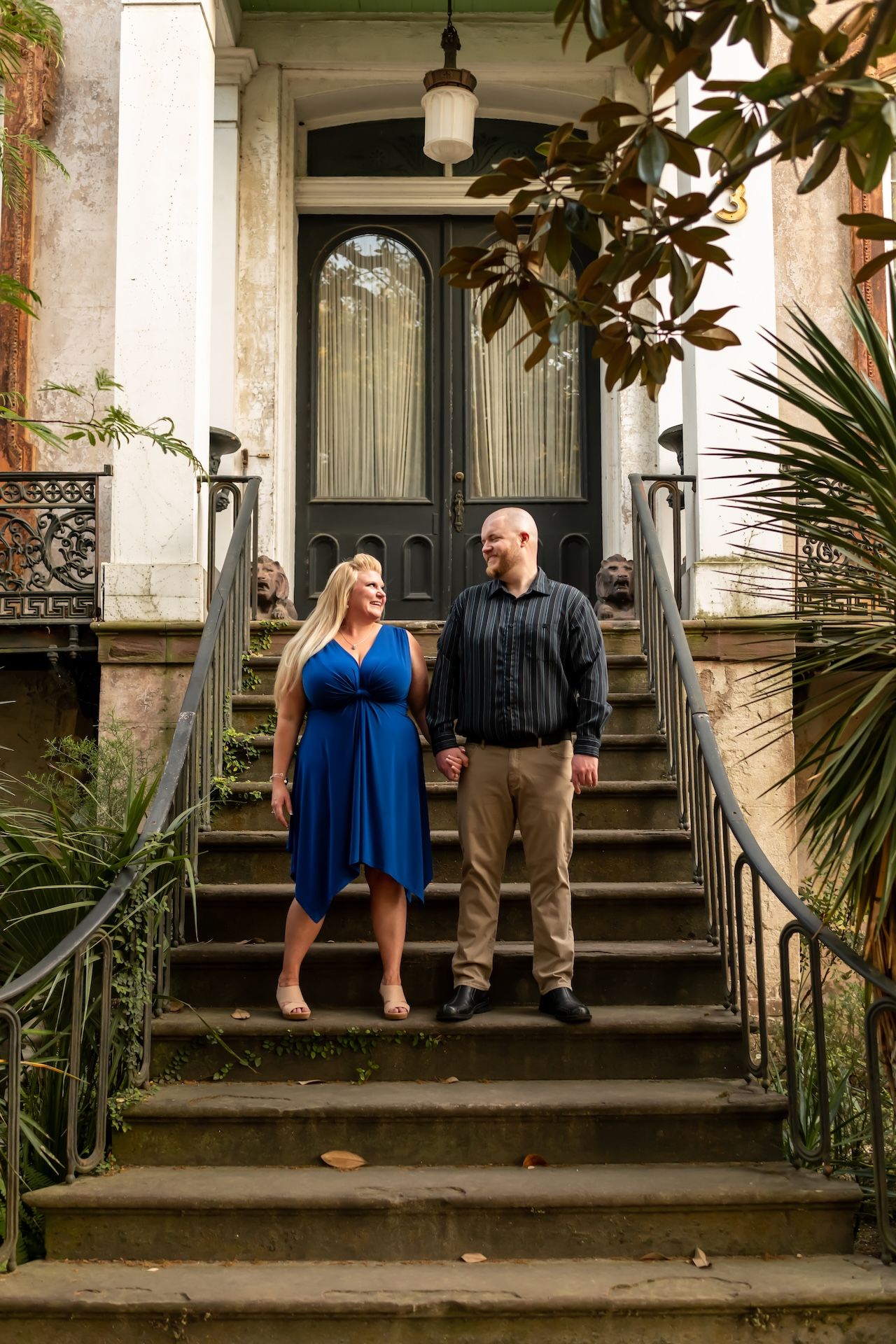 A couple stands together on the steps of a historic southern mansion with magnolia trees and iron railings.