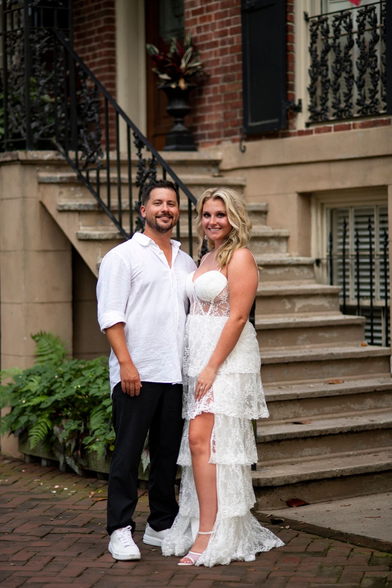 A couple in wedding attire poses on brick steps outside a historic building in an urban setting.