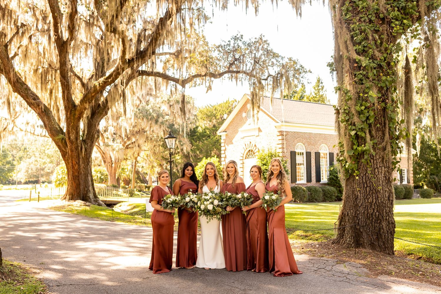 Wedding party in terracotta dresses poses beneath Spanish moss draped oak trees at elegant southern venue in sunlight.