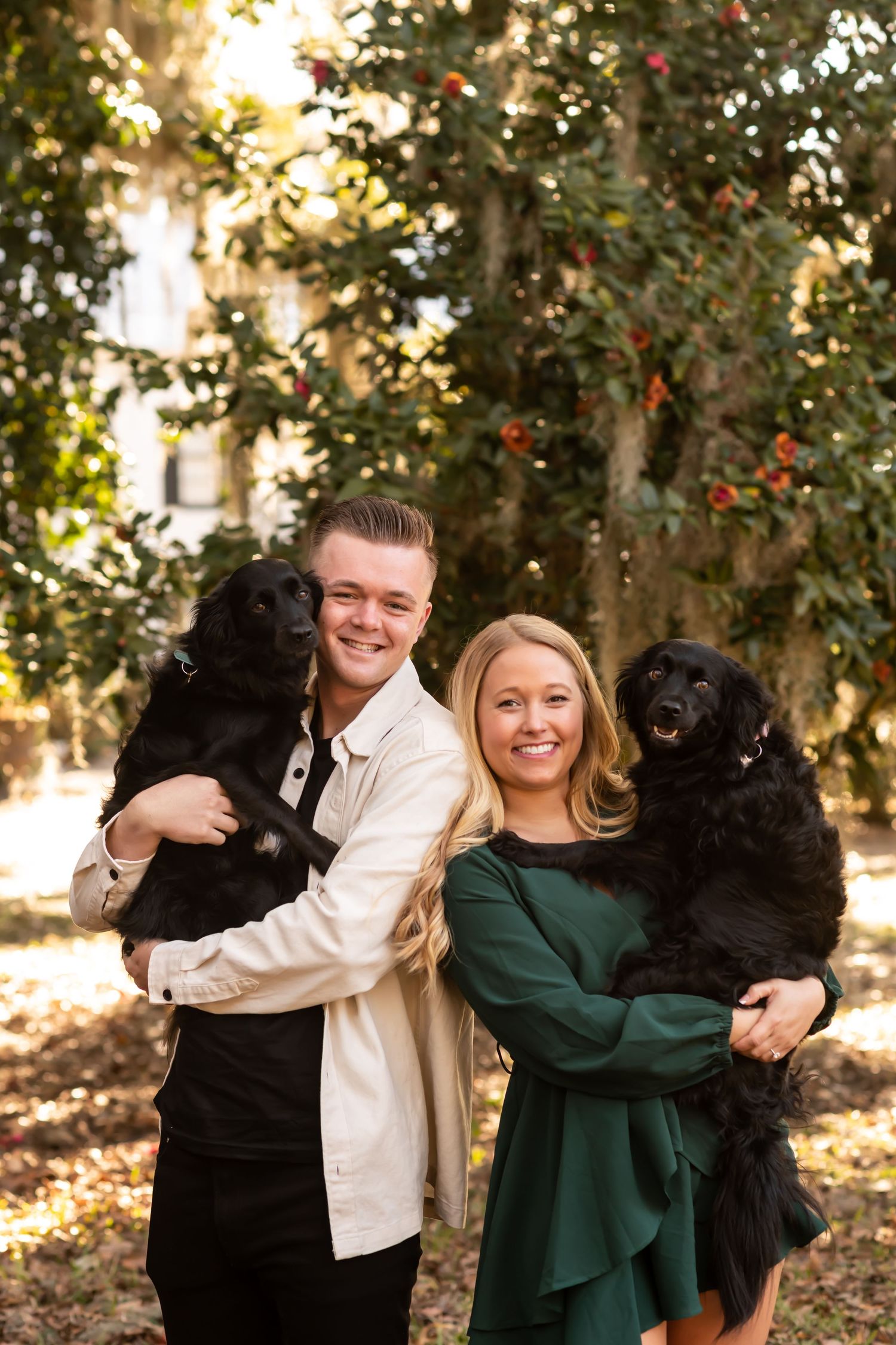 A couple wearing white and green outfits poses outdoors with their two black dogs in a sunlit forest setting.