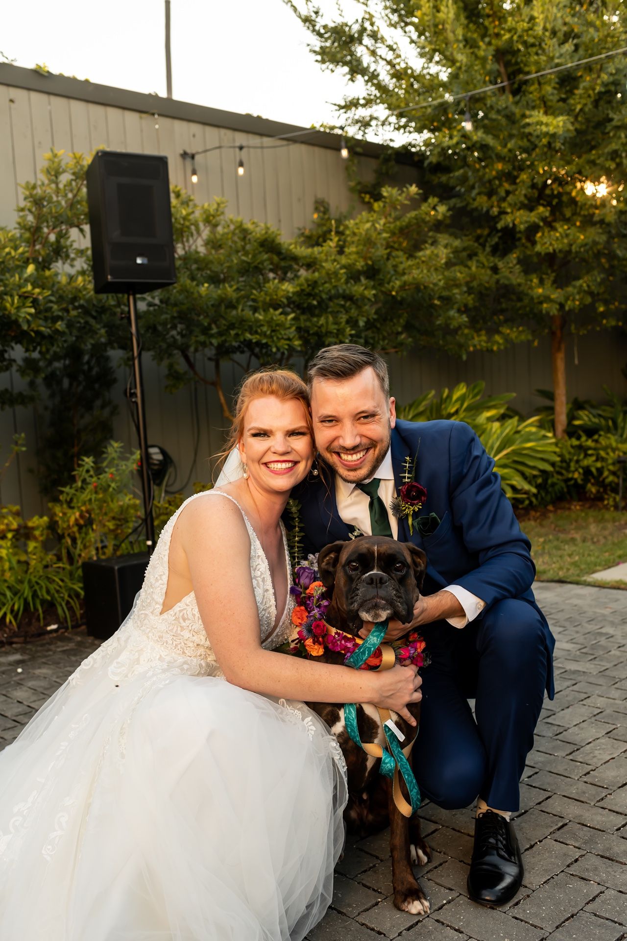 A couple in wedding attire poses with their black dog on a stone paved patio near a building and trees.
