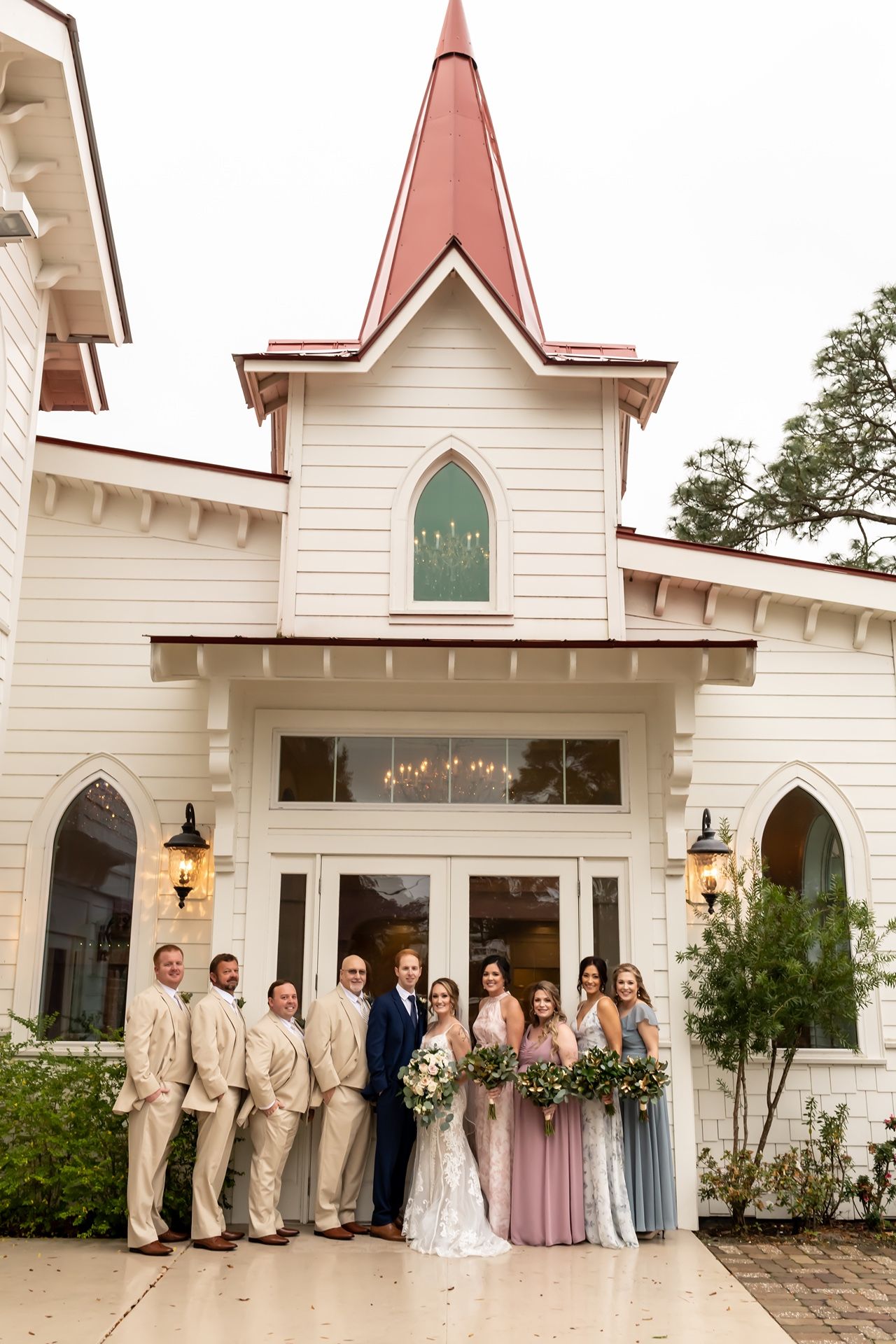Wedding party poses in front of a charming white chapel with a pink spire and Gothic arched windows.