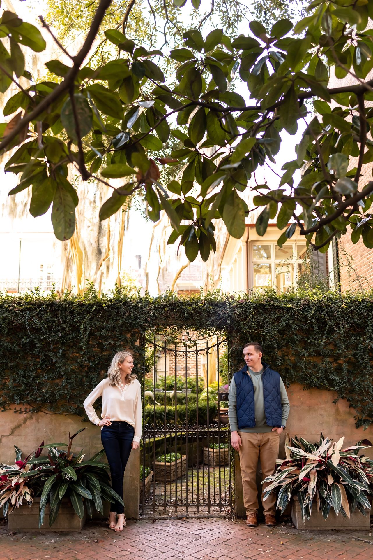 A couple stands beside a decorative wrought iron gate surrounded by lush greenery and magnolia trees.