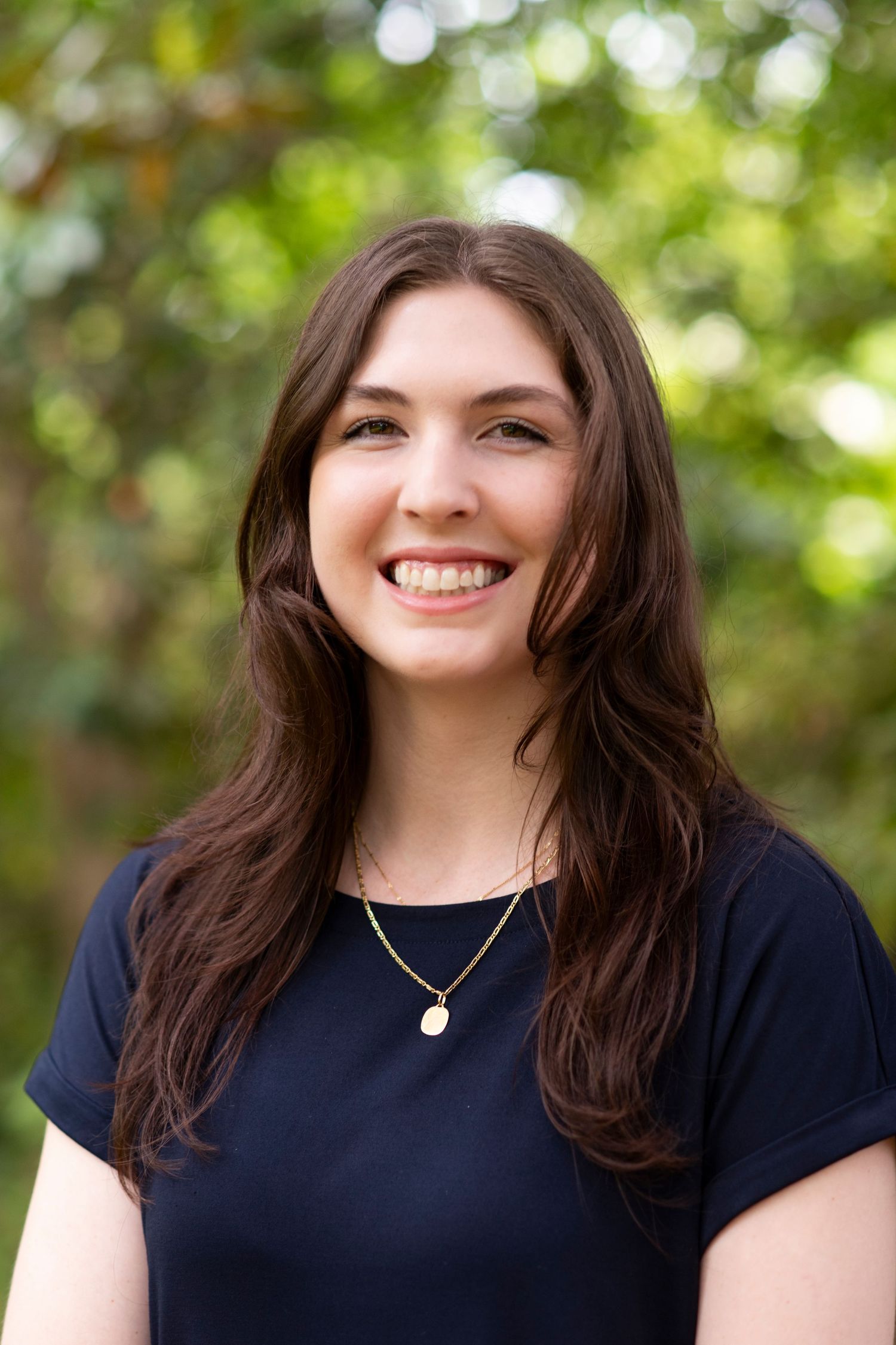 A woman in a dark navy blue top smiles warmly against a soft, blurred green outdoor background.