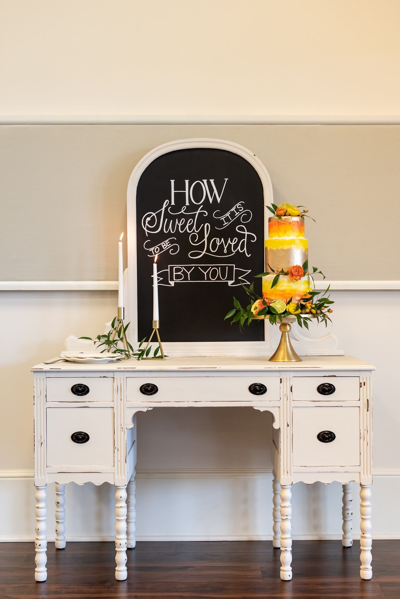 Elegant white vintage vanity with decorative chalkboard sign and yellow floral wedding cake display on hardwood floor.