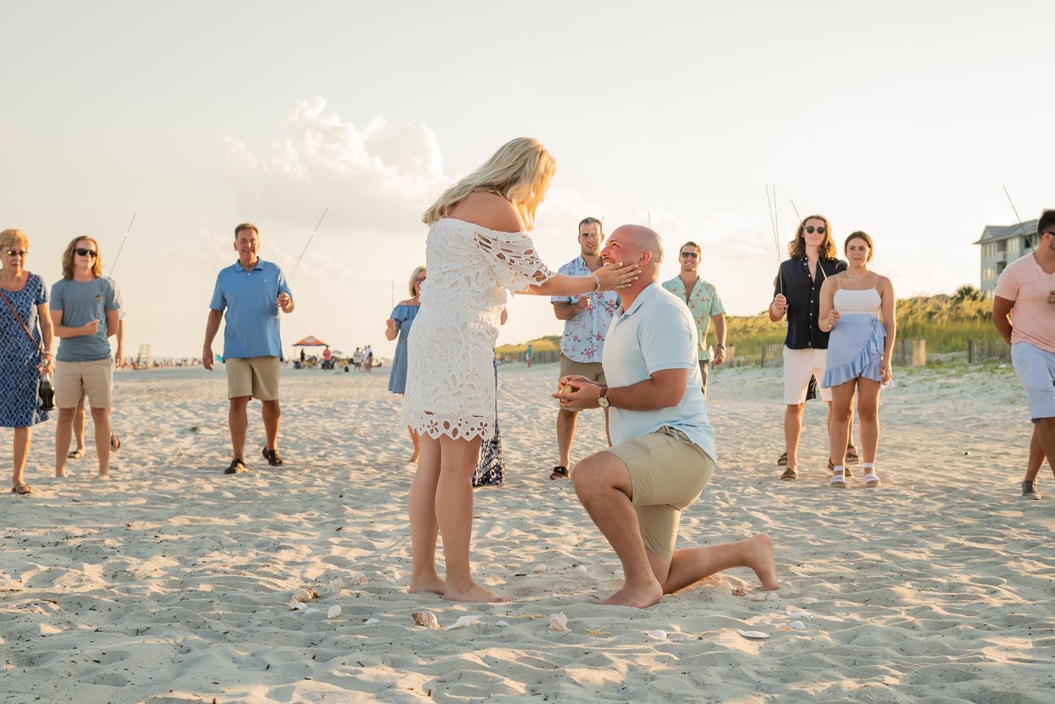 A beachside proposal scene at sunset with onlookers watching a special moment unfold on the sandy shore.