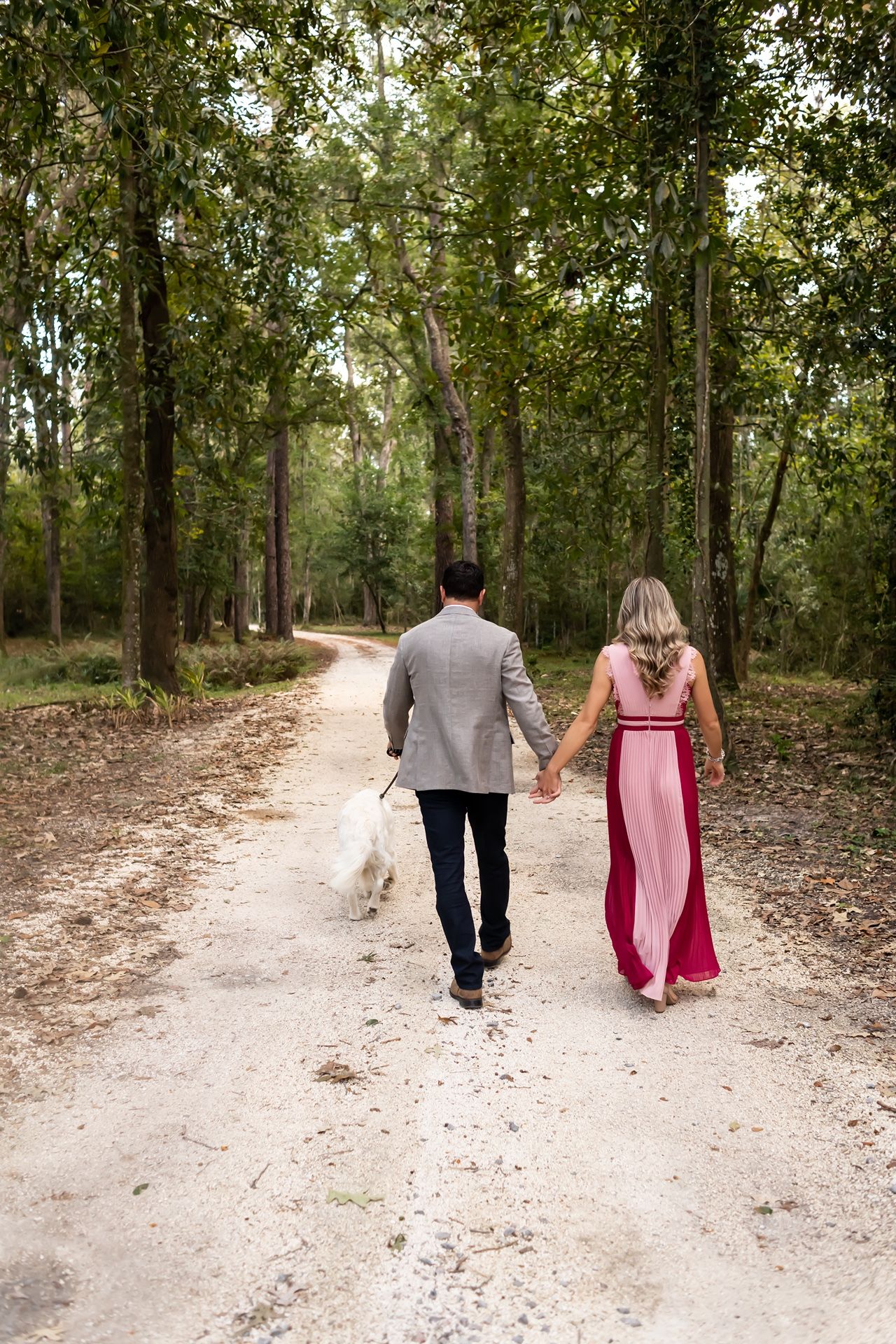 A couple in formal wear walks their dog down a scenic dirt path through a lush wooded forest.