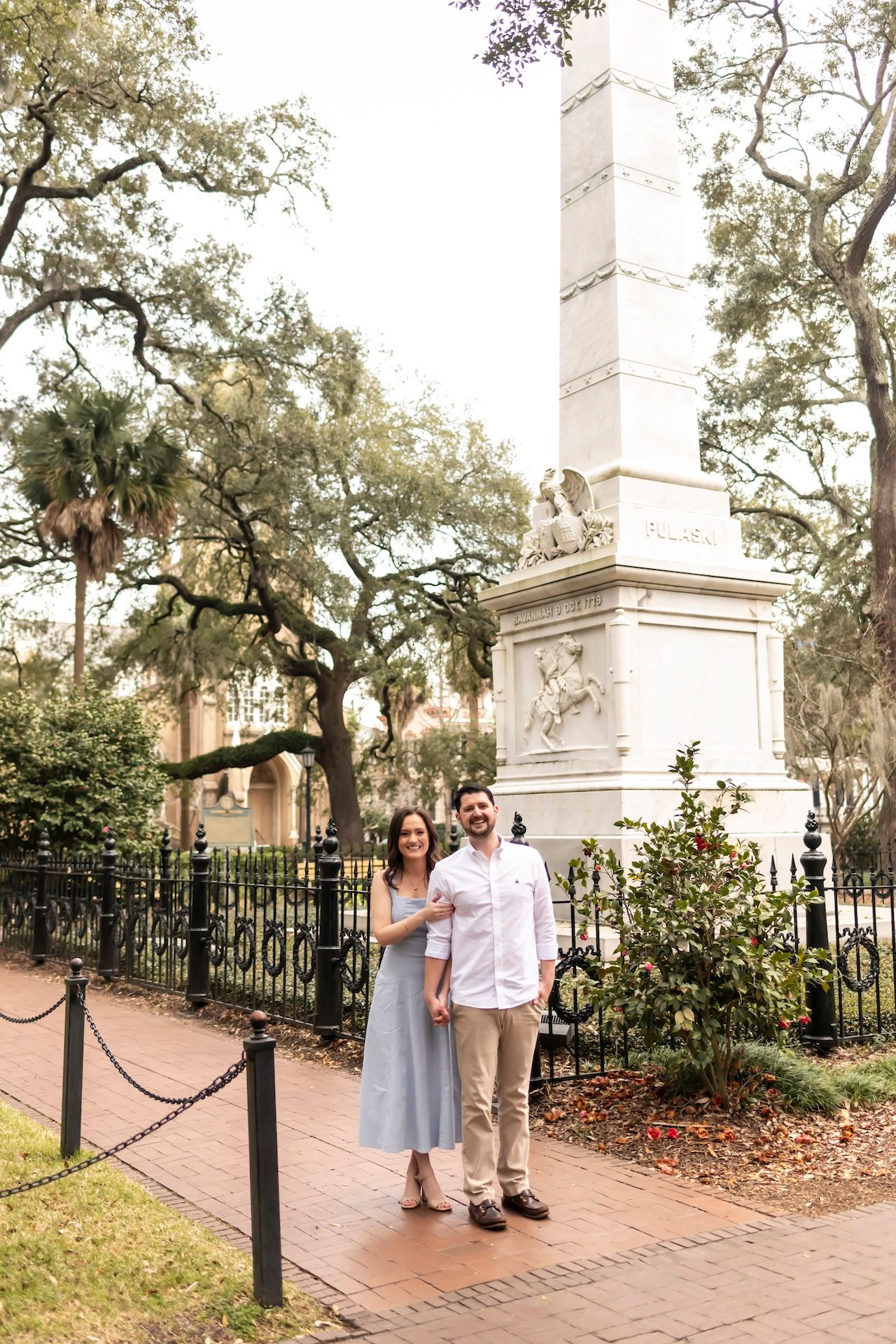 A couple stands together along a brick pathway under Spanish moss draped trees near white stone monuments.