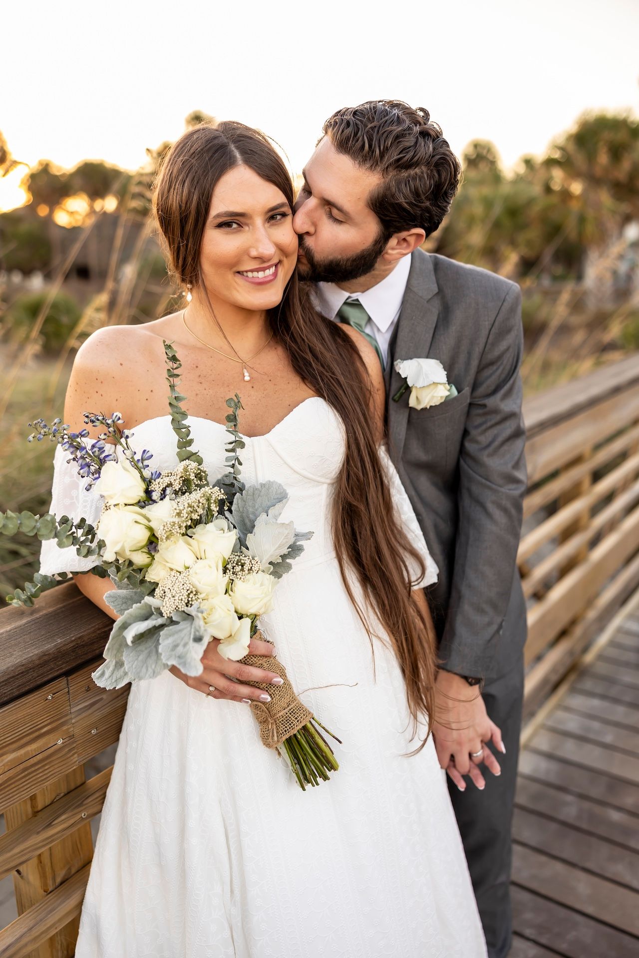 A couple on their wedding day sharing an intimate moment on a wooden bridge at sunset with a white floral bouquet.