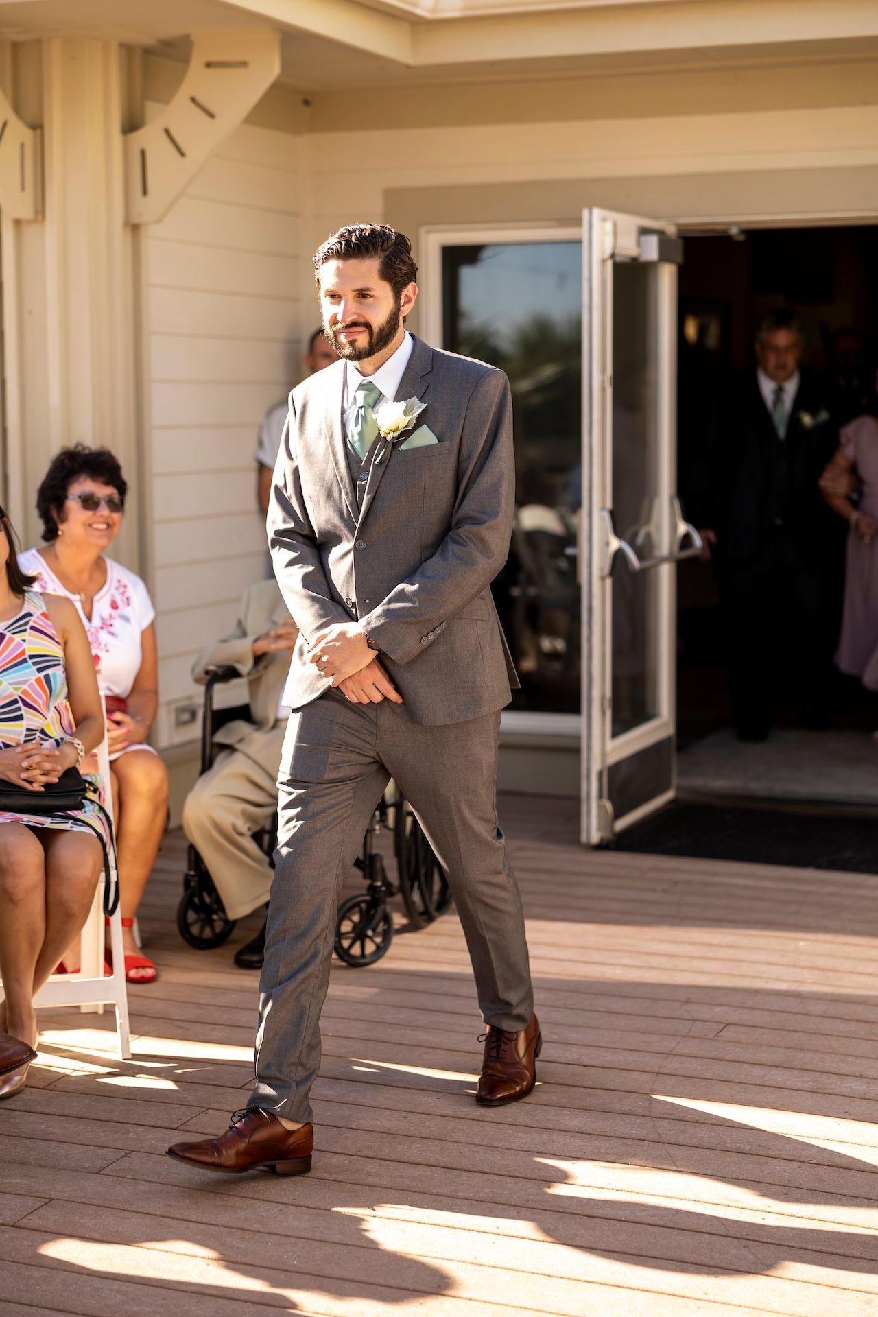Wedding participant in gray suit and boutonniere walking across sunny outdoor deck.