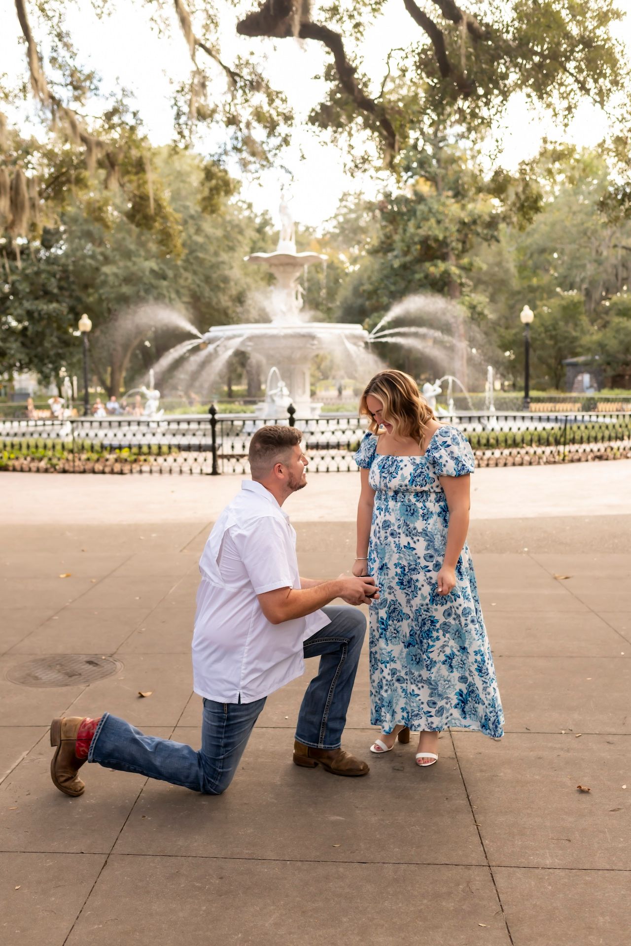 A romantic proposal scene at a scenic fountain in a park with trees and gardens in the background.