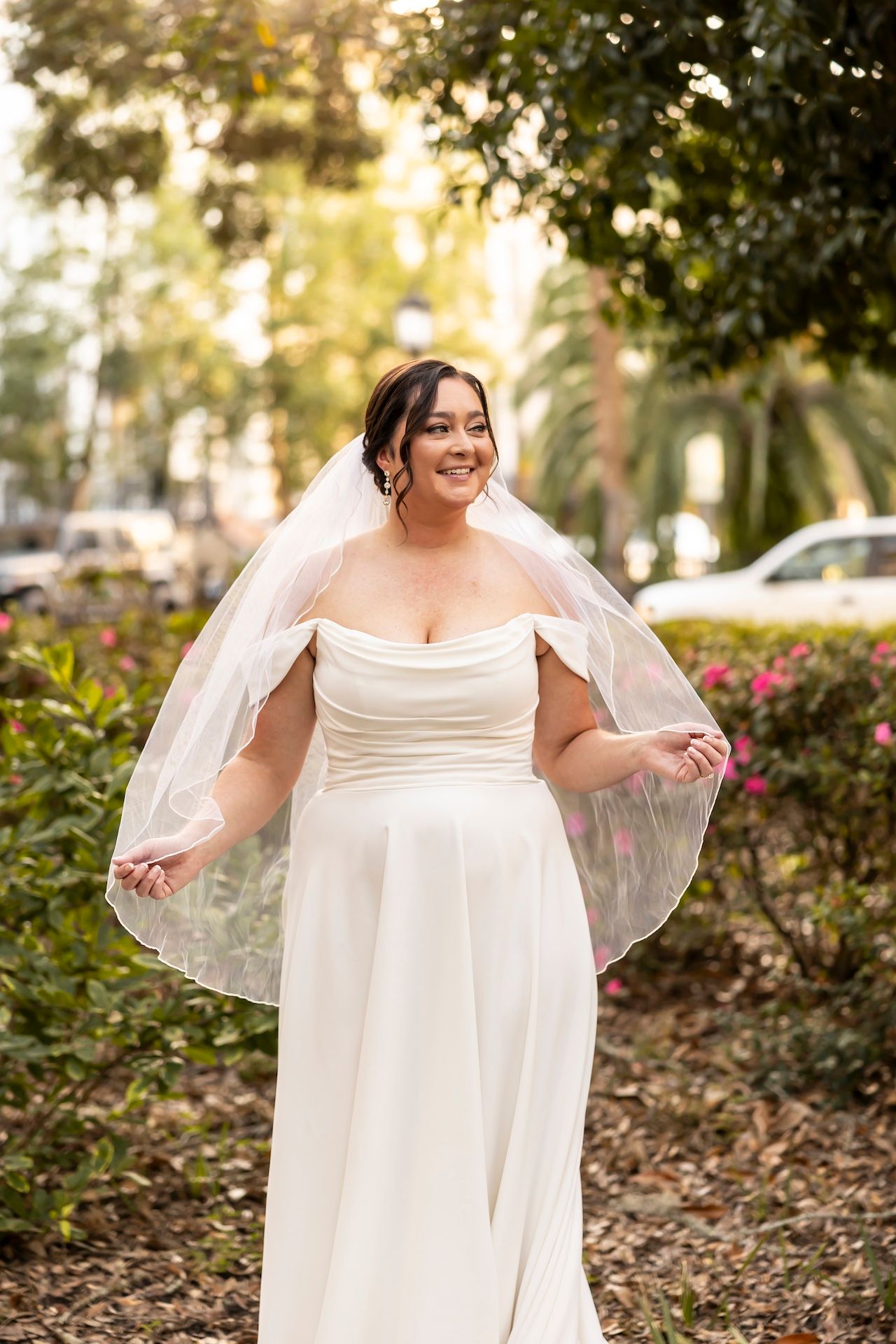 The bride wears a strapless white wedding gown with a flowing veil in a garden setting at sunset.