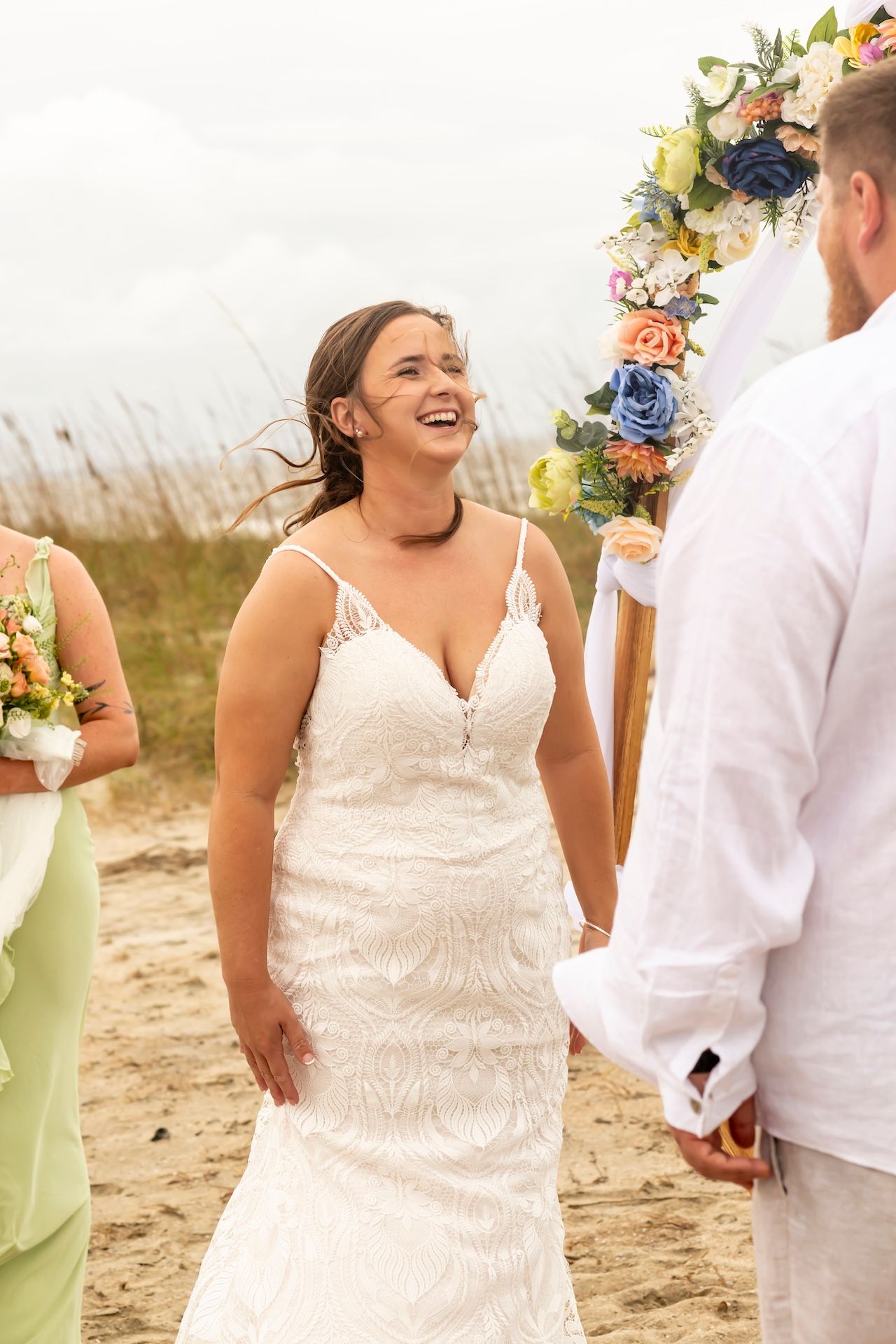 Joyous bride laughs during beach wedding ceremony under floral arch with soft lighting and coastal backdrop.