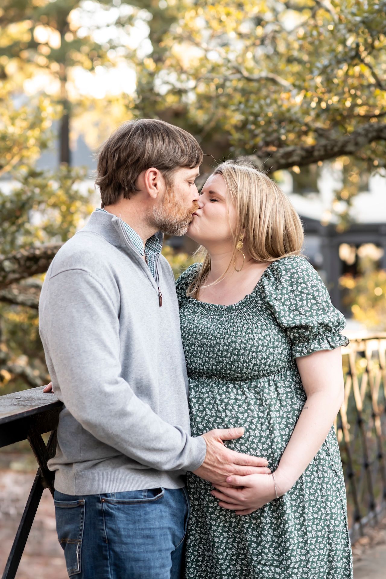 Couple in romantic kiss while cradling pregnant belly on outdoor bridge.