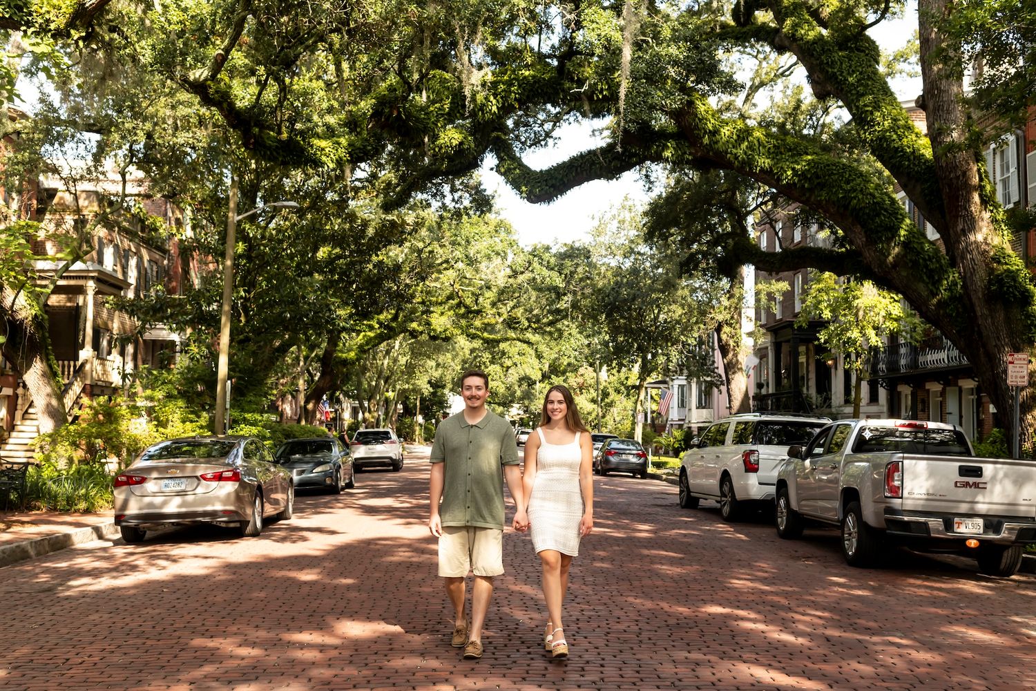 Tree-lined cobblestone street with parked cars and overhanging oak branches in a residential neighborhood.