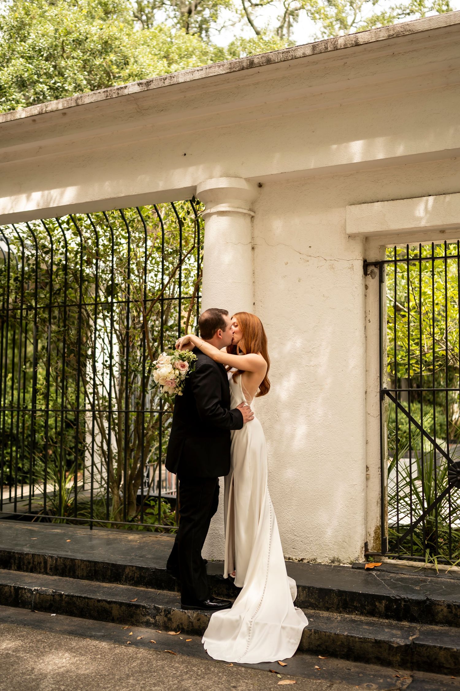 A couple embrace and kiss on steps of a white columned porch with wrought iron railings and lush greenery in summer.