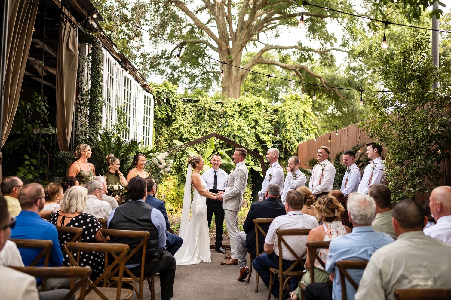 Outdoor wedding ceremony takes place in a lush garden setting with wooden chairs and natural decor.