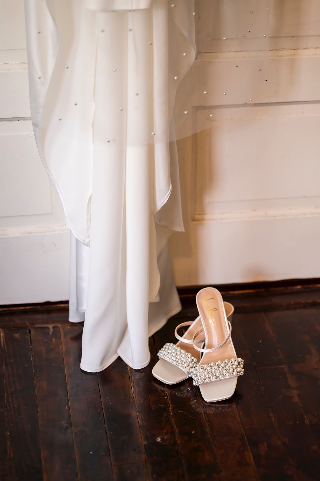 A series of bridal shoes peeking out from under a white wedding dress hem against a dark wooden floor.