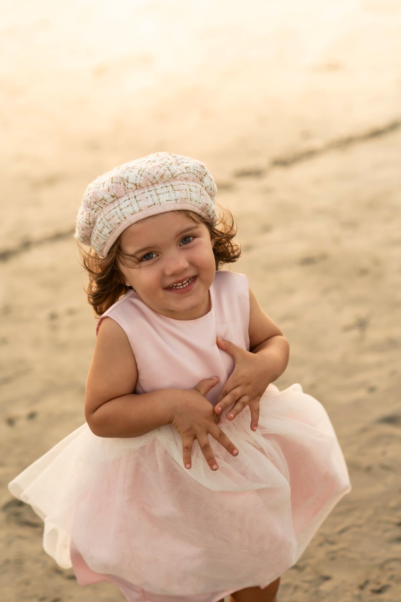 A young child in a pink tulle dress and patterned cap smiles while playing on sandy ground.