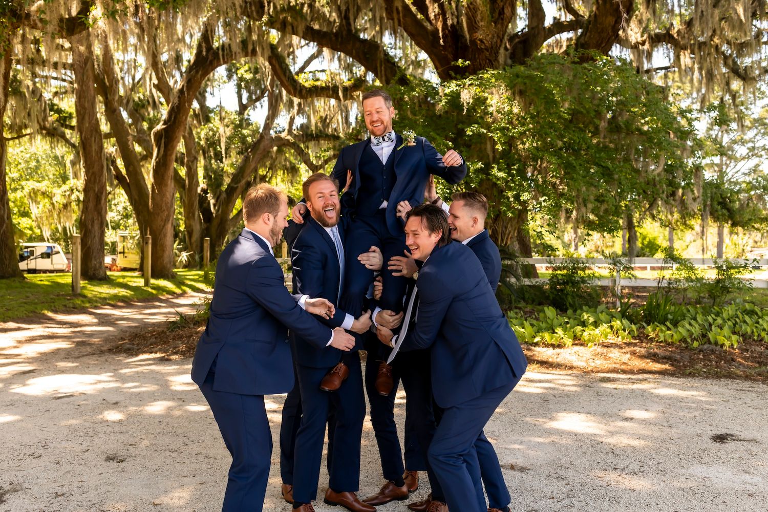 A group in matching navy suits laugh together on a path under Spanish moss draped oak trees in a garden setting.