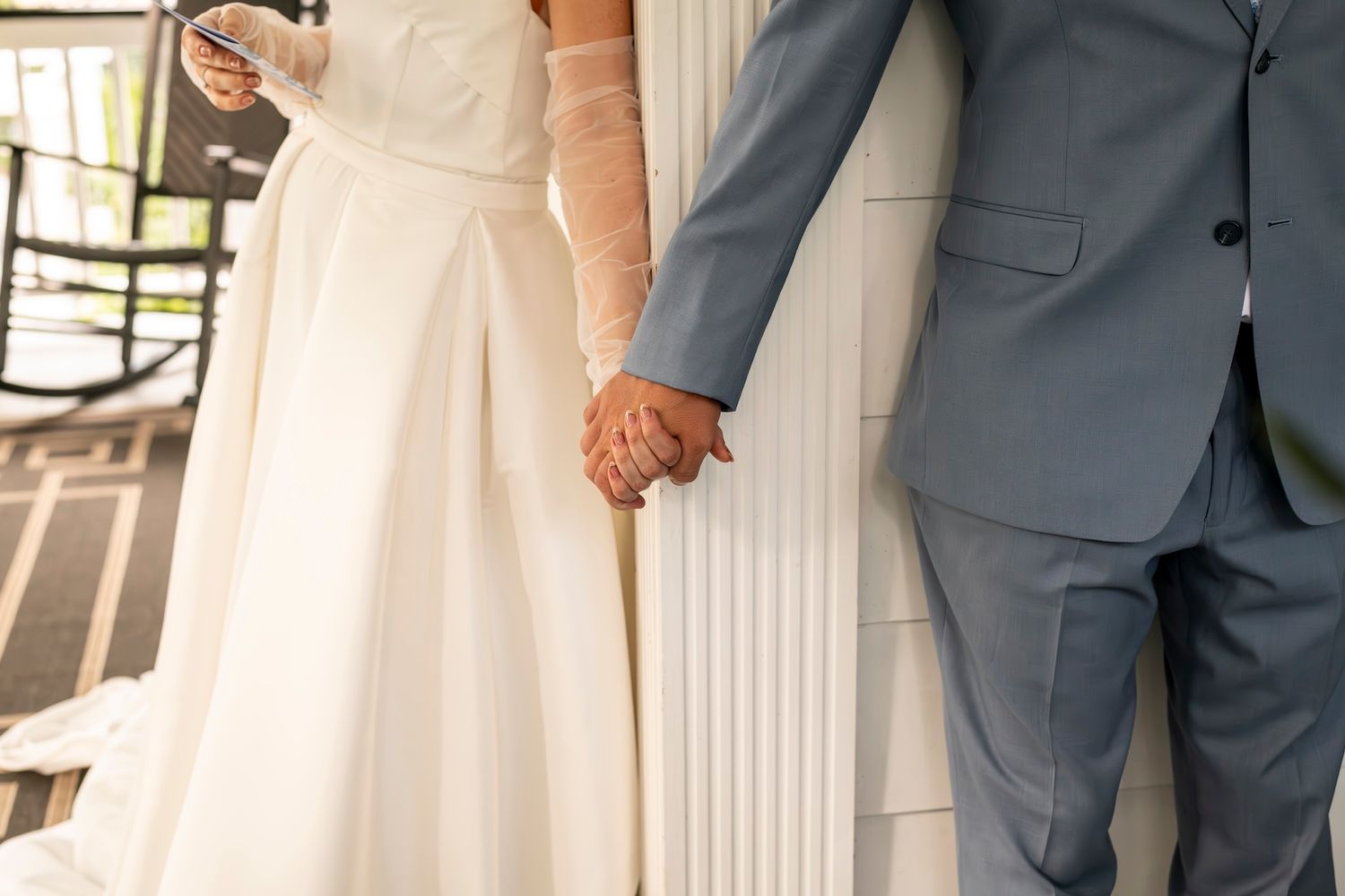 Close-up view of a bride in white dress and groom in gray suit holding hands during wedding ceremony.