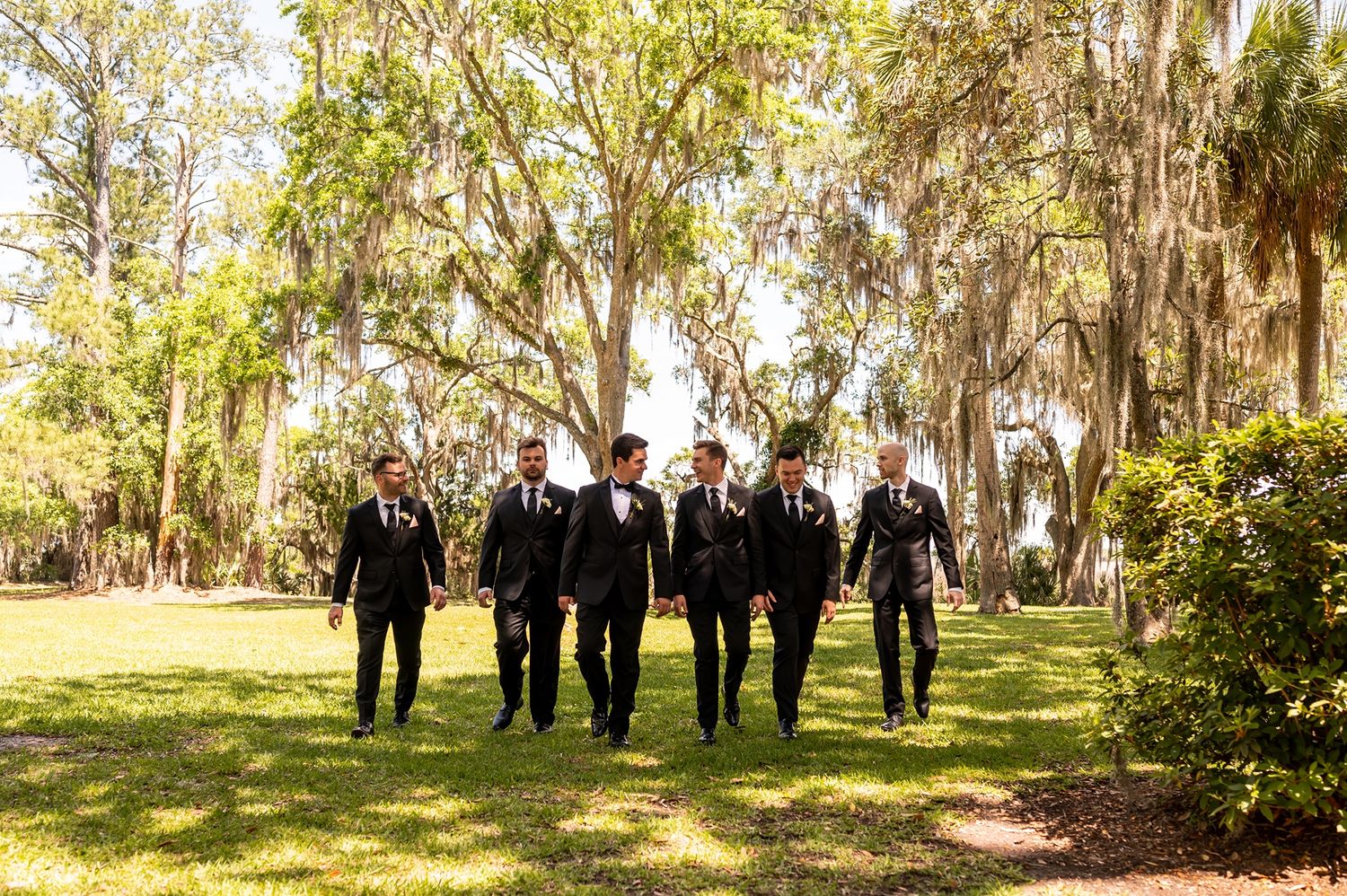 A group walks together outdoors on a path through a scenic garden with Spanish moss hanging from trees.