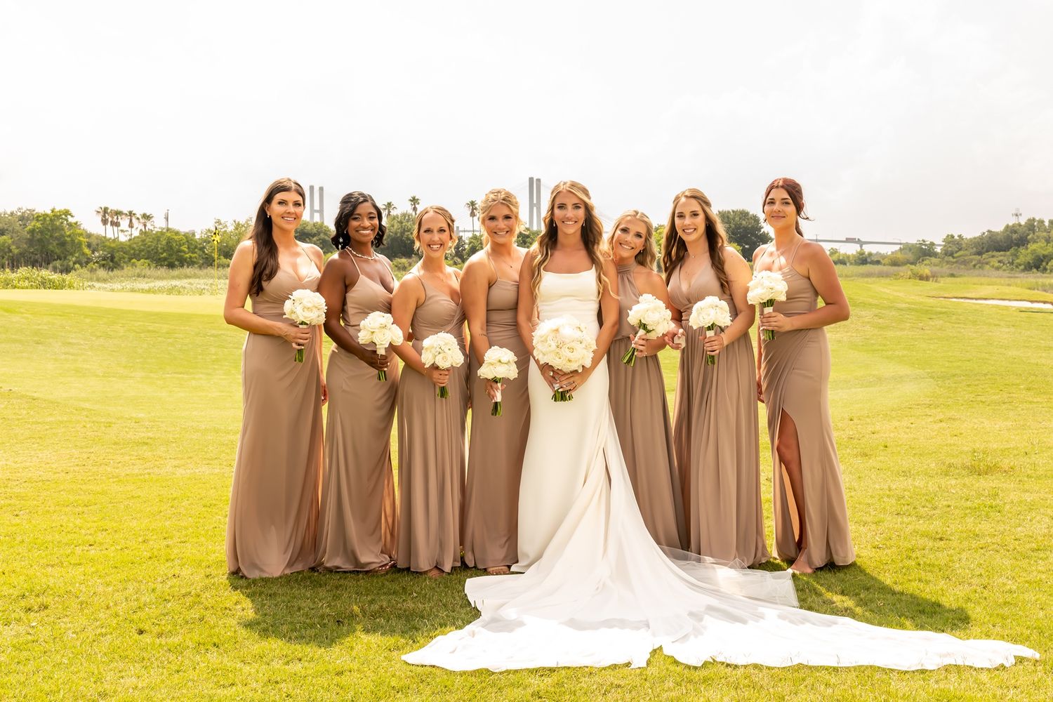 A wedding party in beige bridesmaid dresses and a white bridal gown pose together on a grassy outdoor field.