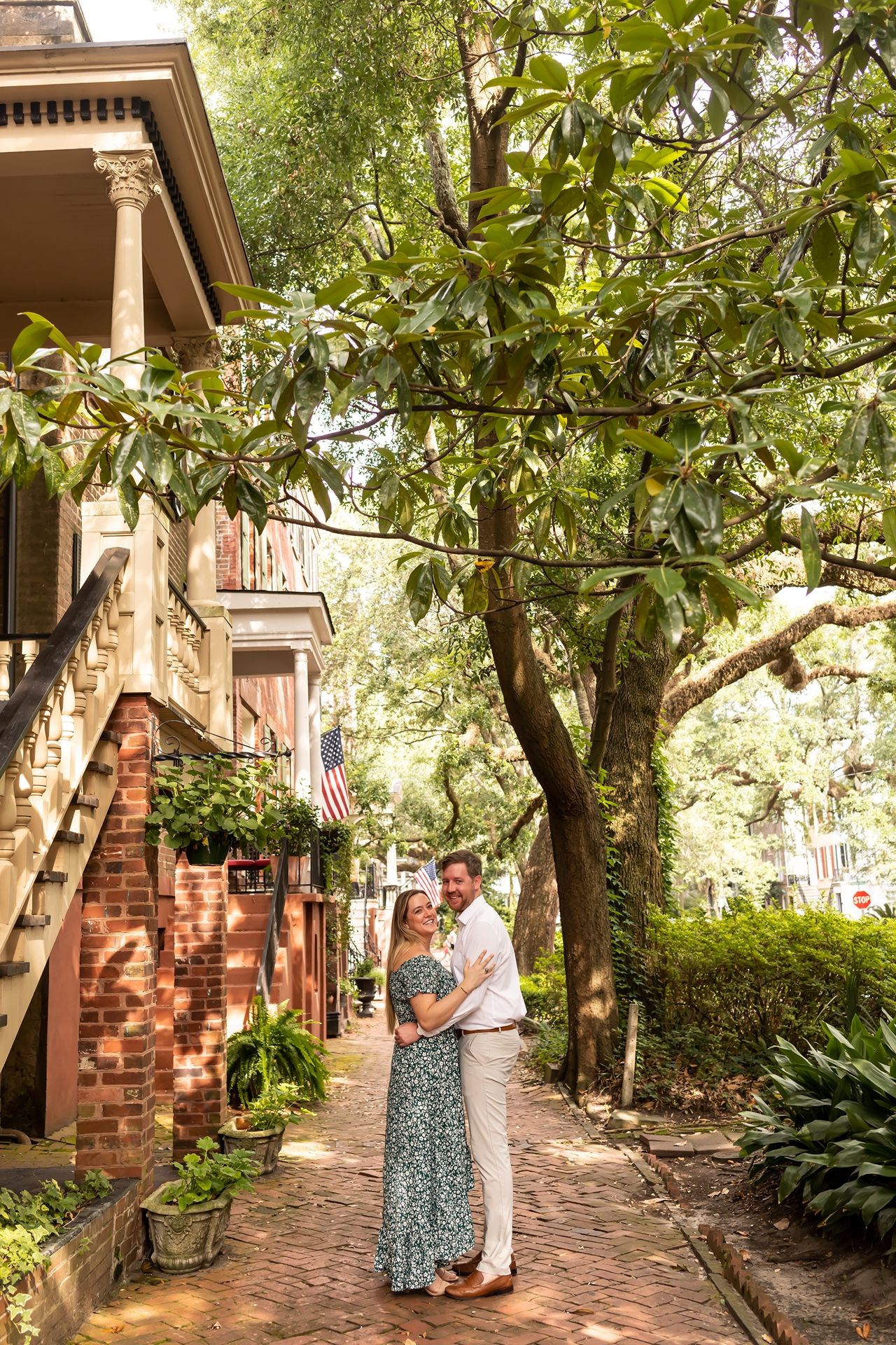 A romantic couple shares intimate moments along a charming brick pathway lined with lush greenery and trees.