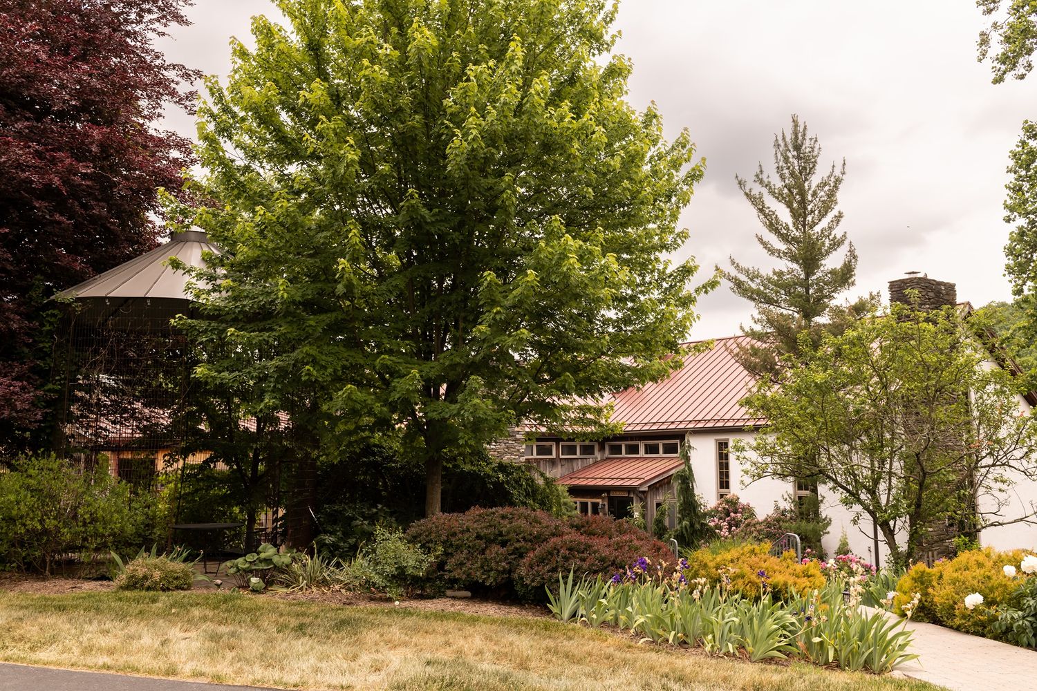 Landscaped garden with colorful flowers surrounds a brown house with umbrella patio seating under mature trees.