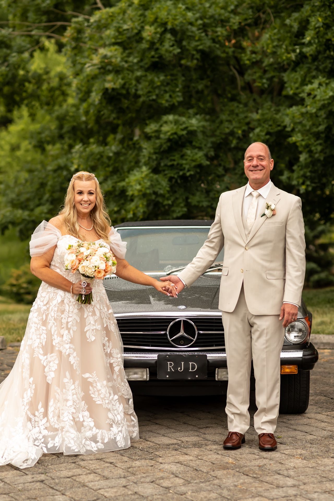 A wedding couple poses in front of a classic Mercedes-Benz car, the bride in lace and groom in beige suit holding hands.