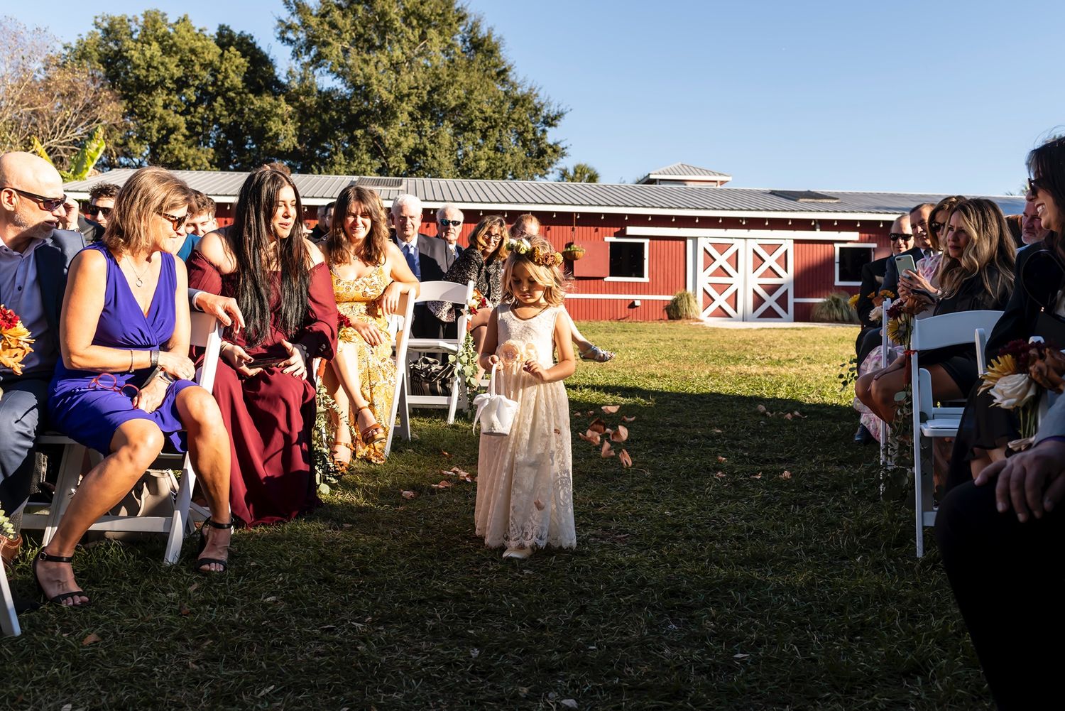 Child in a white dress scatters flower petals down a grass aisle during an outdoor wedding ceremony at a rustic barn venue.