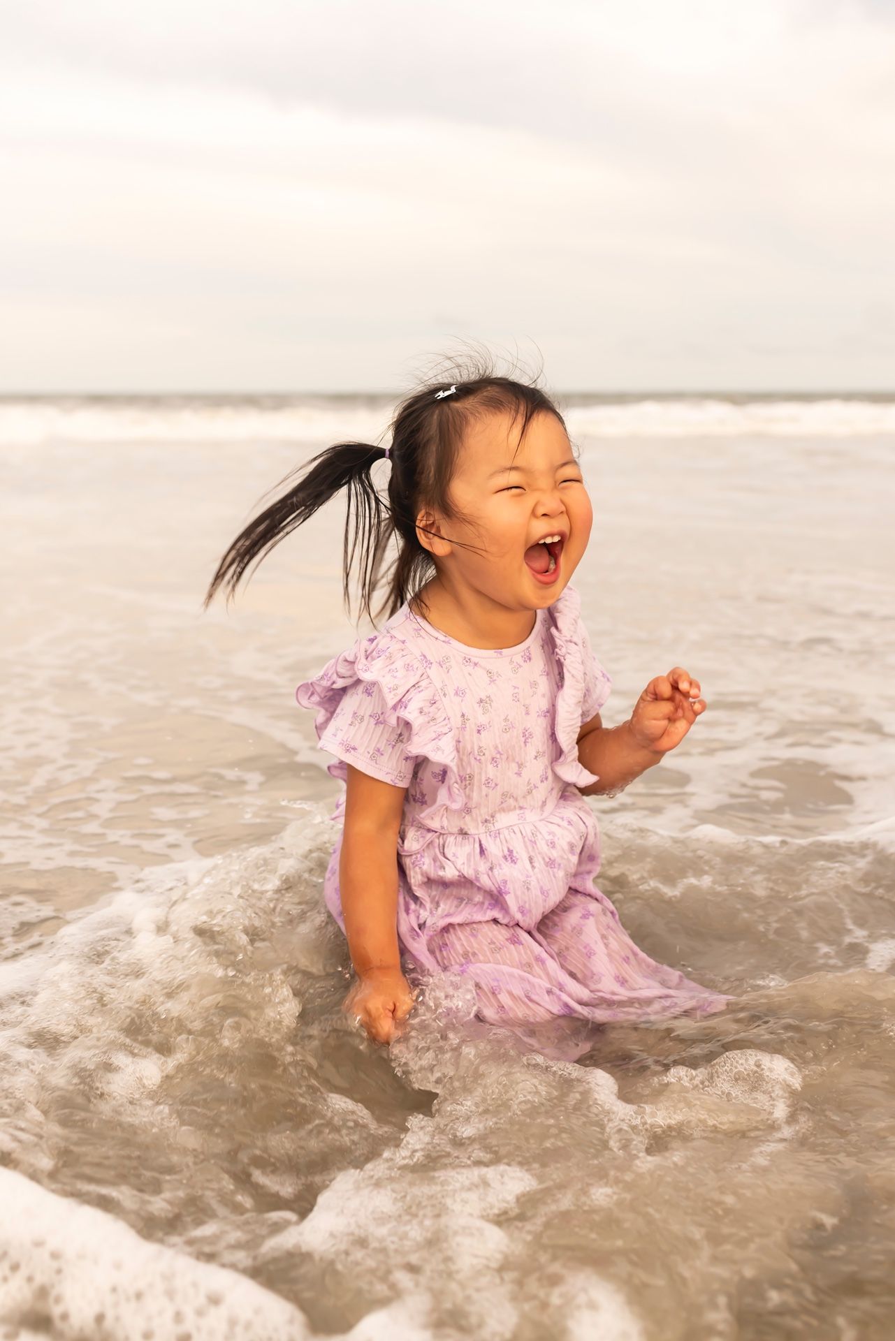 A young child in pink dress laughs joyfully while playing in ocean waves at the beach.