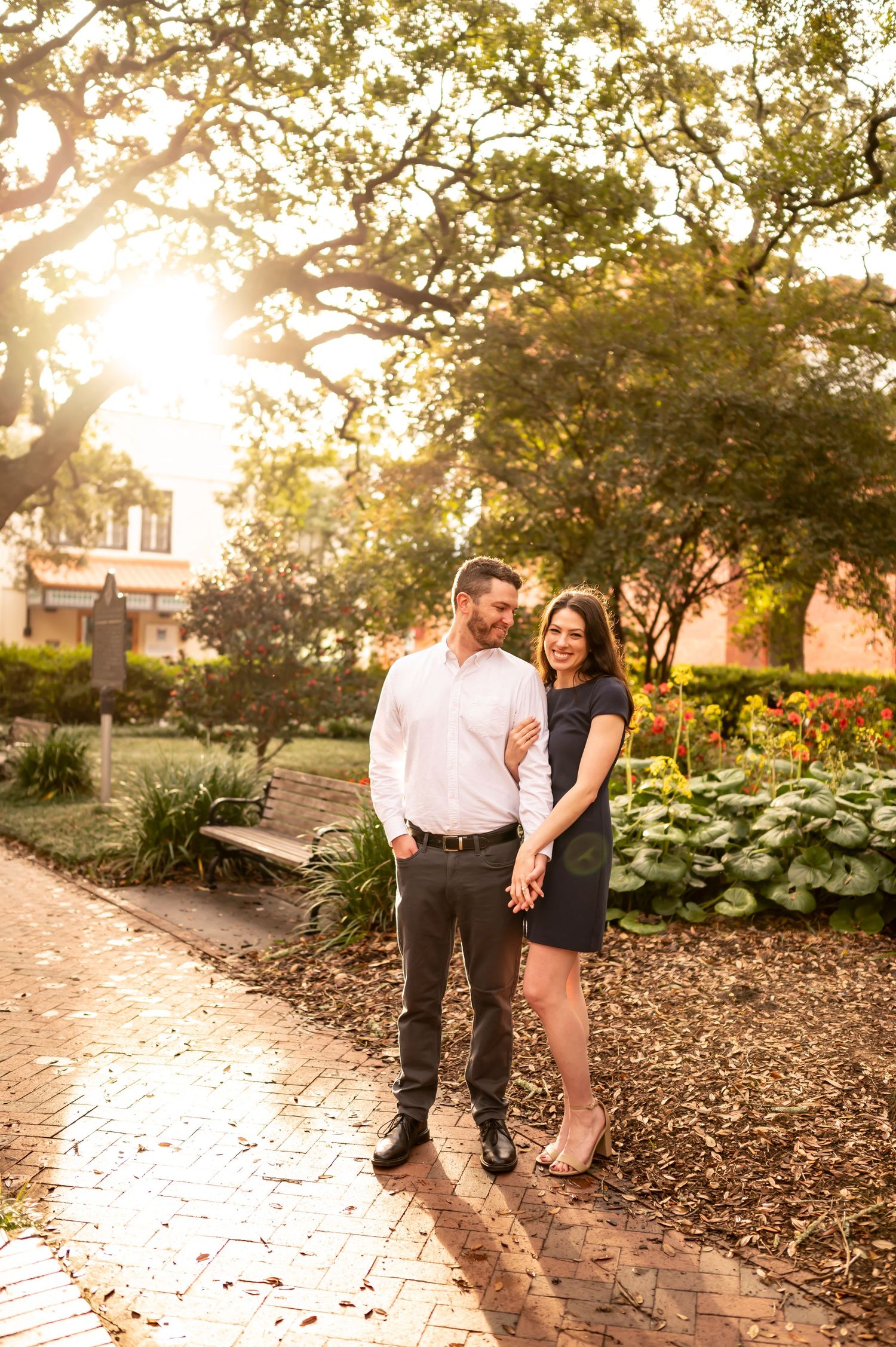 A couple stands together on a sunlit brick path in a garden with golden light filtering through trees at sunset.