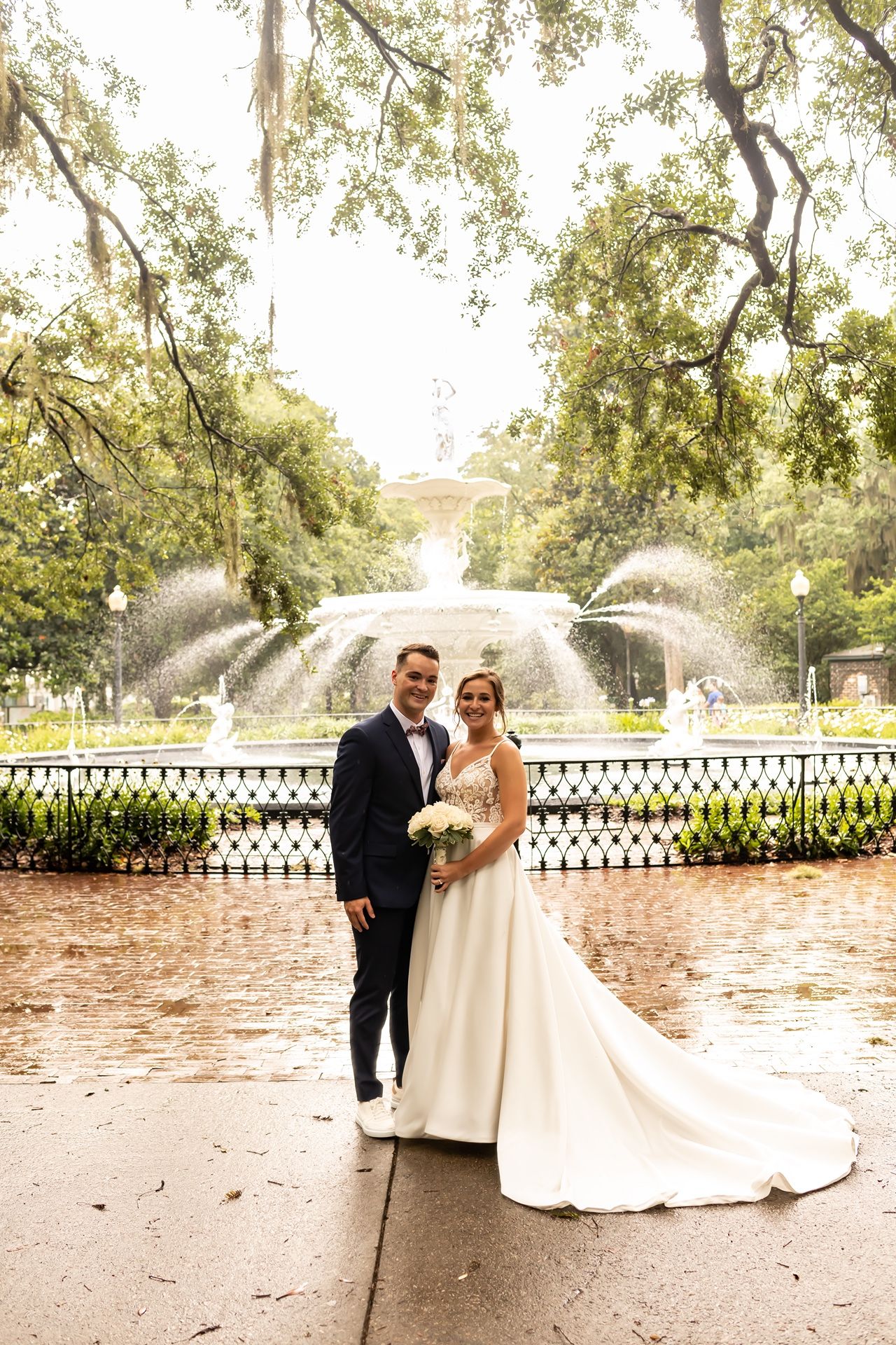 A wedding couple poses in front of a beautiful fountain in a garden setting with Spanish moss draped trees.