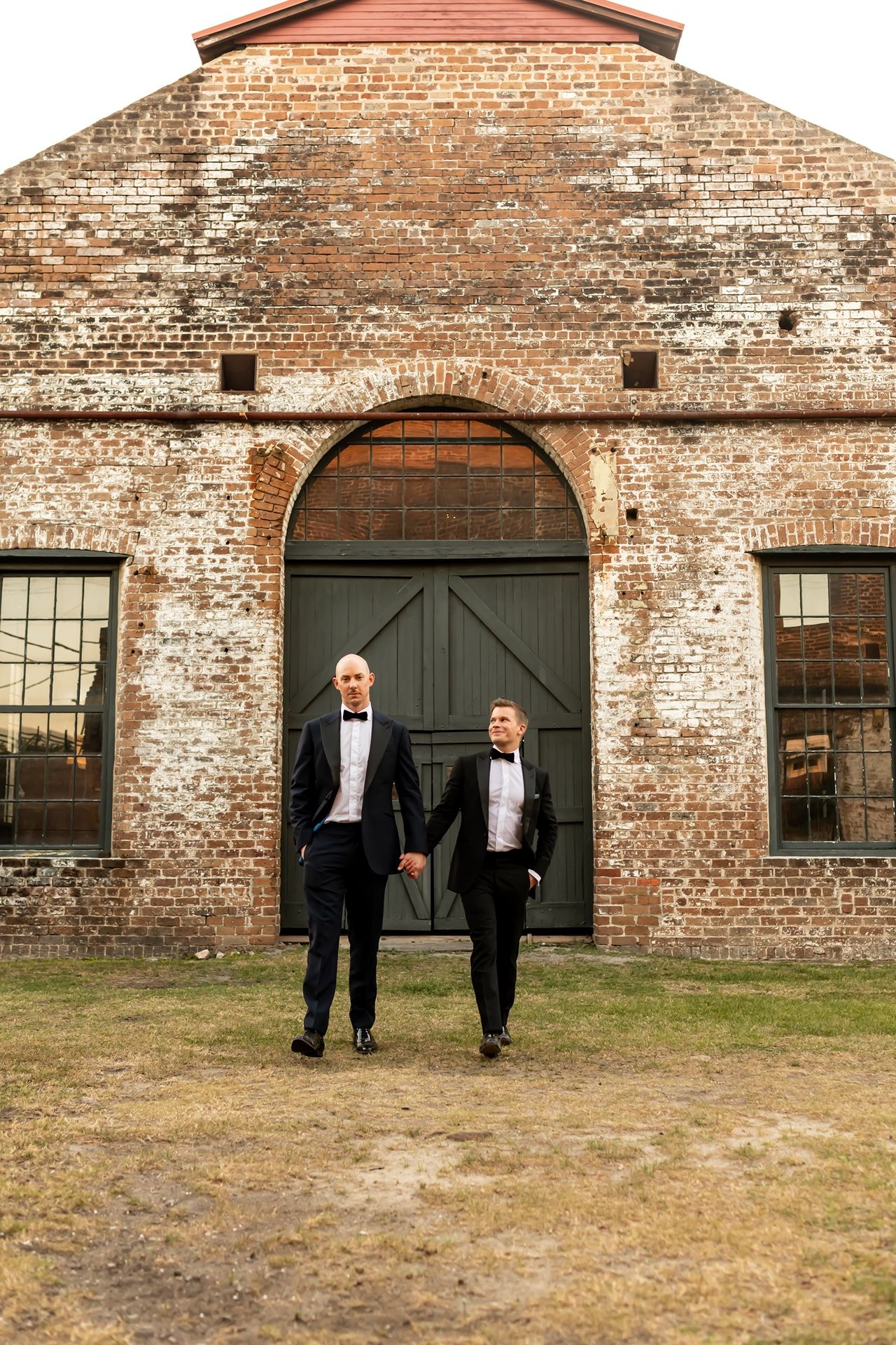 Two people in classic tuxedos walk hand in hand in front of a rustic brick industrial building with arched doorway.