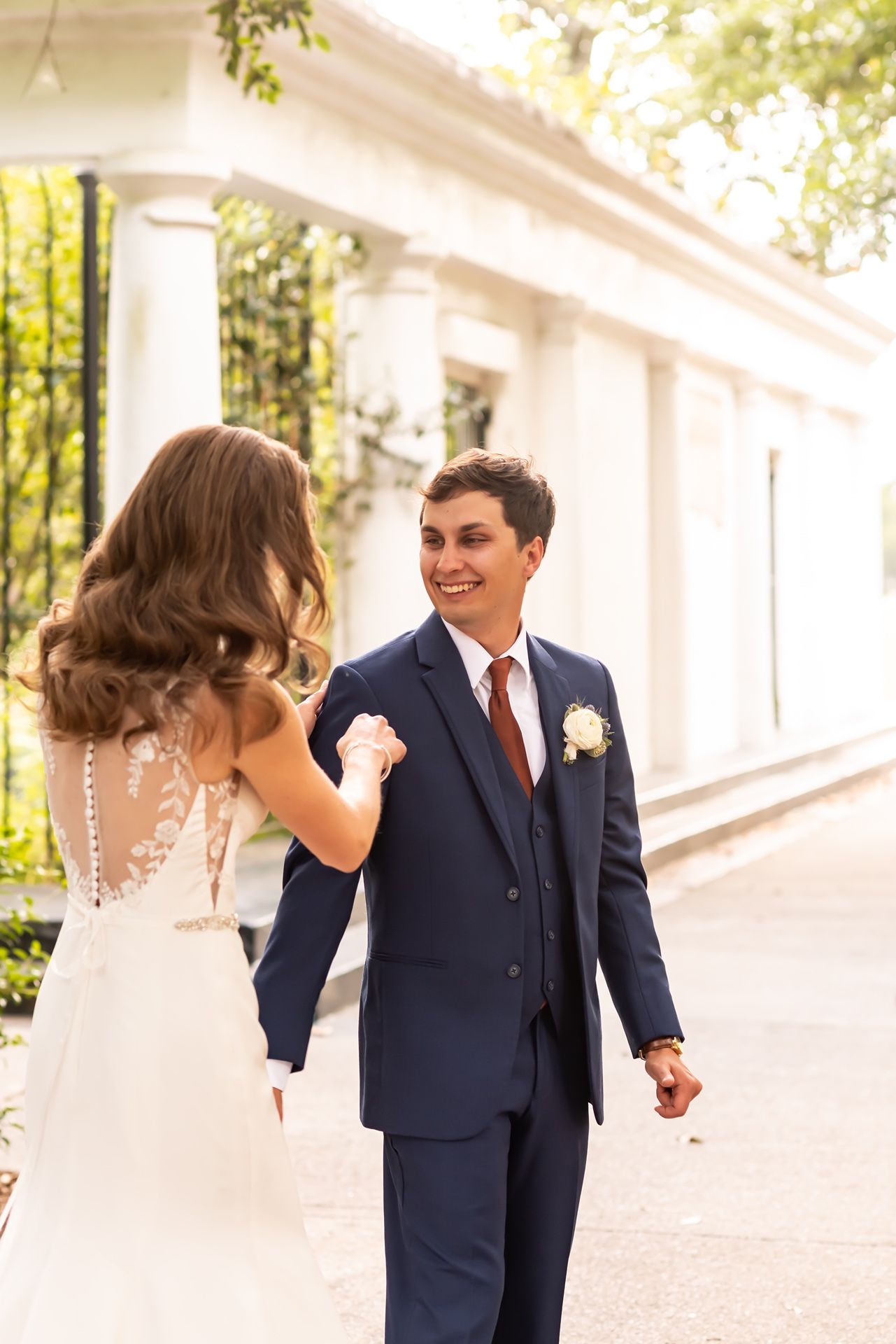 Romantic wedding couple walks hand in hand along a white columned walkway in elegant navy suit and flowing dress.
